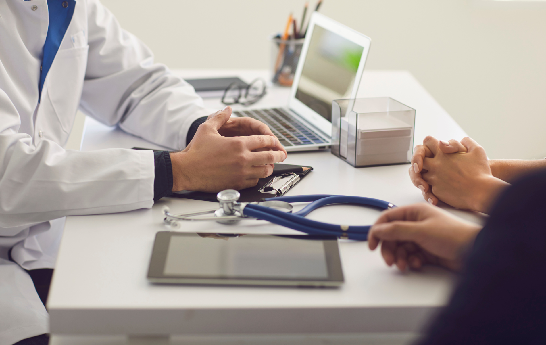 Doctor and patient discussing at a clinic desk with a laptop and stethoscope nearby.