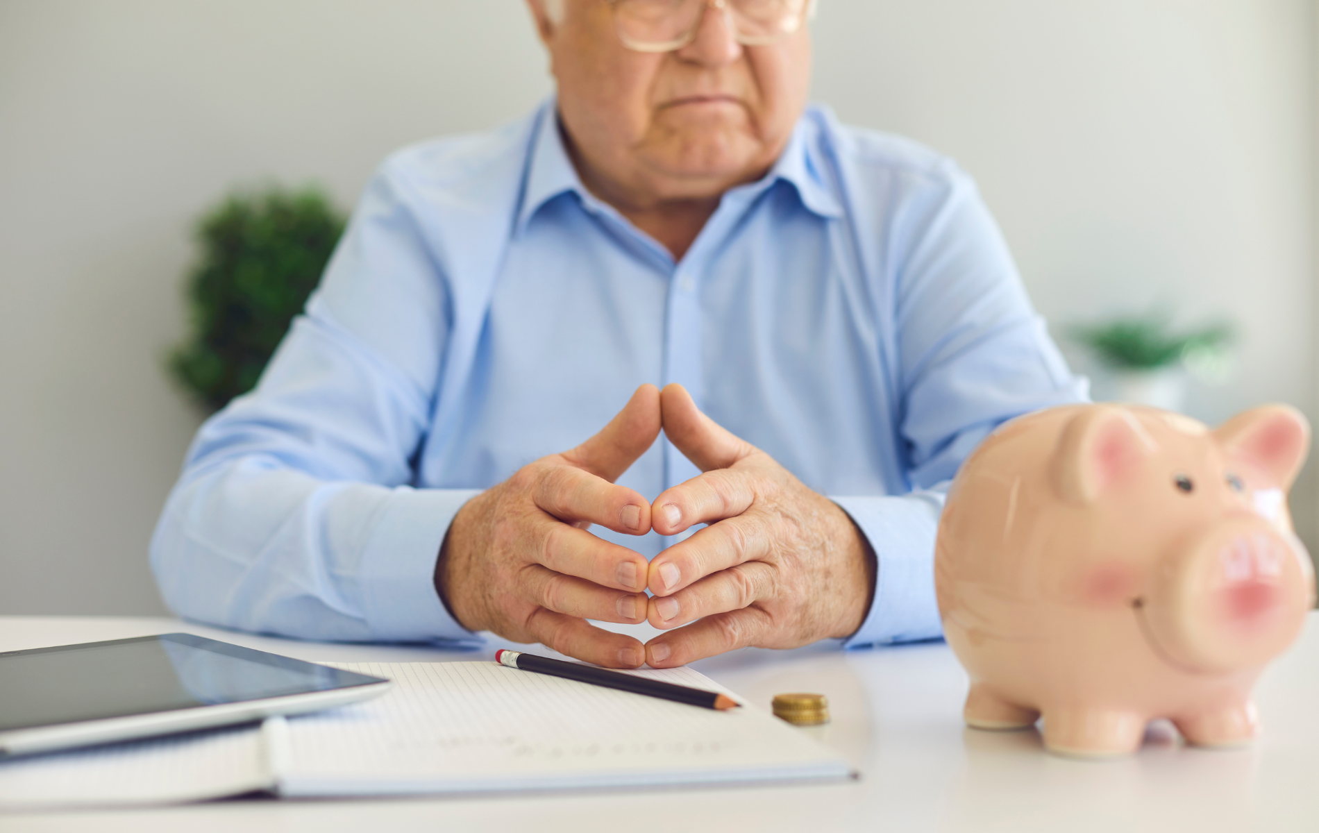 Person in a blue shirt with clasped hands beside a piggy bank, coins, and laptop on a desk