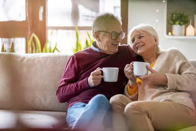 Two people on a couch smiling and holding white mugs in a cozy living room
