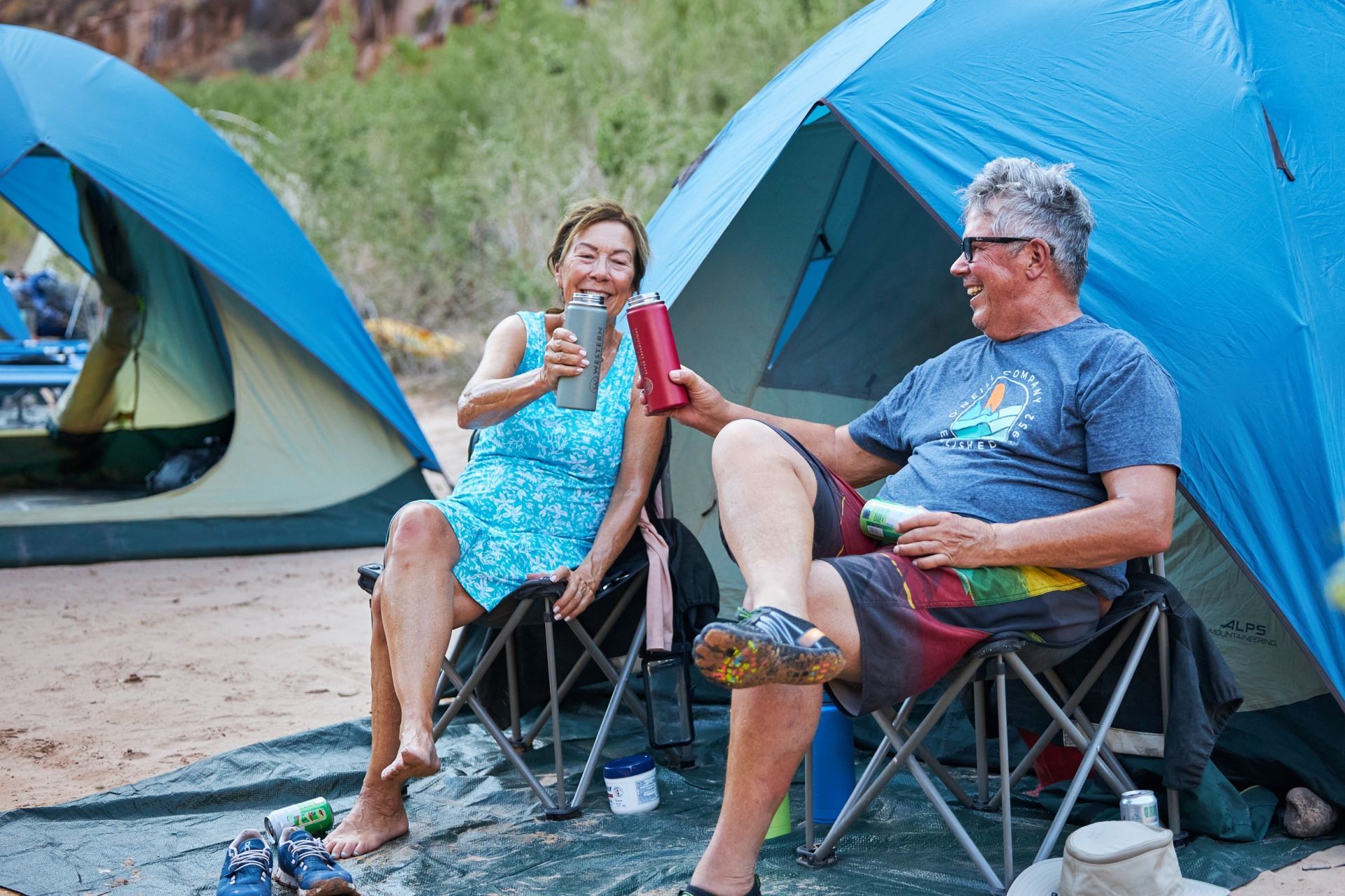 Two campers relax in folding chairs beside their river-camp tents