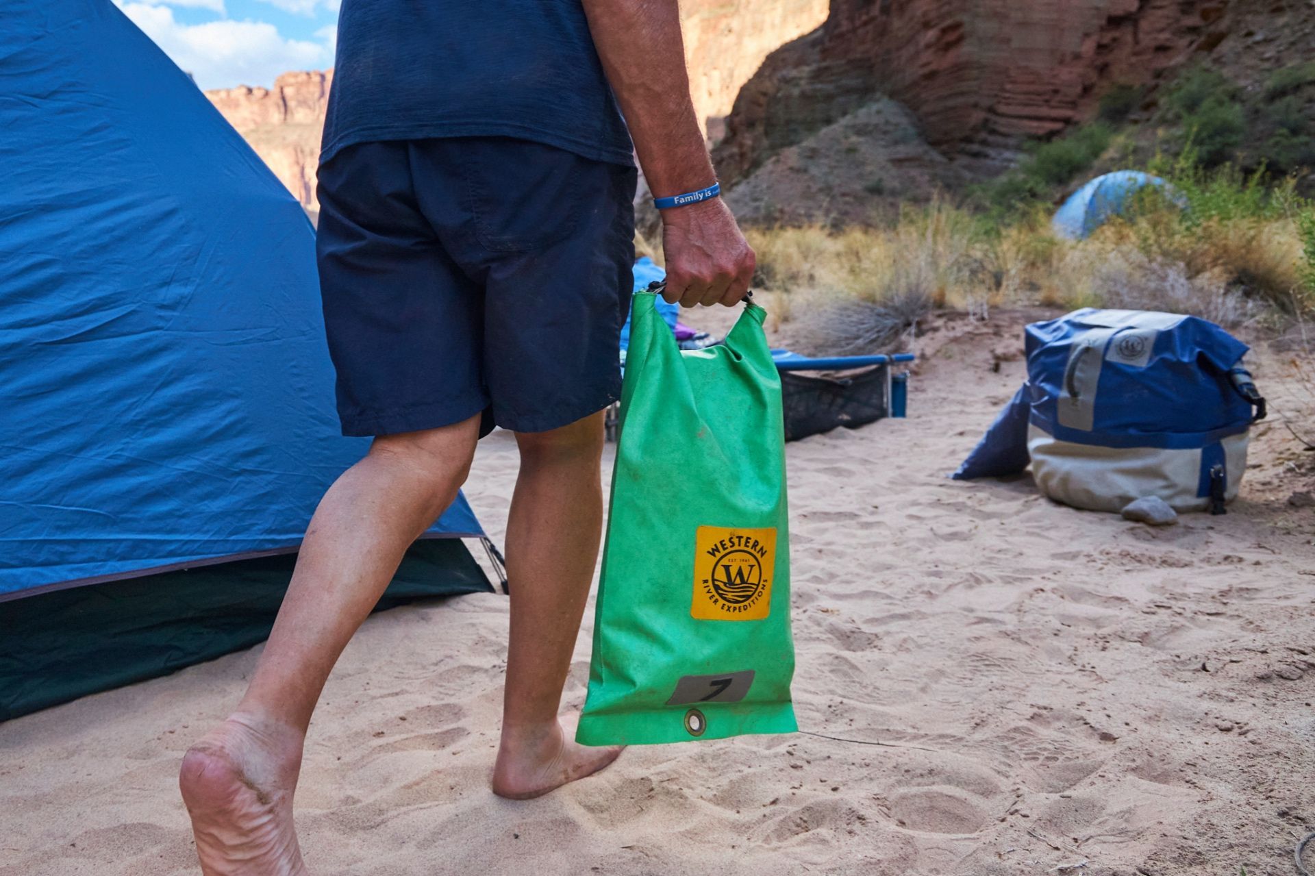 Camper carrying a green dry bag through a riverside camp