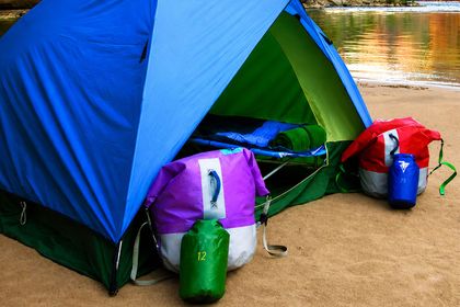 A blue tent at rivers edge with purple and red dry bags out front.