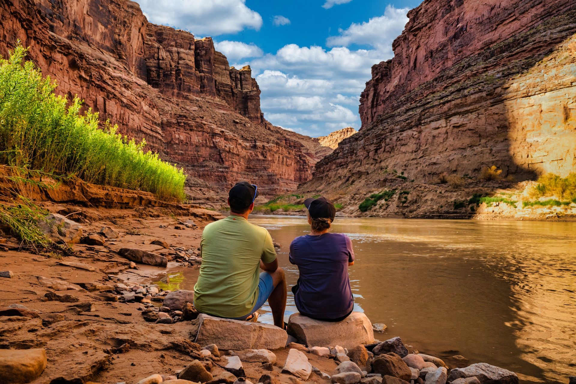 A couple sits at the river's edge, looking downstream at tall canyon walls as the sun reflects off the water. 
