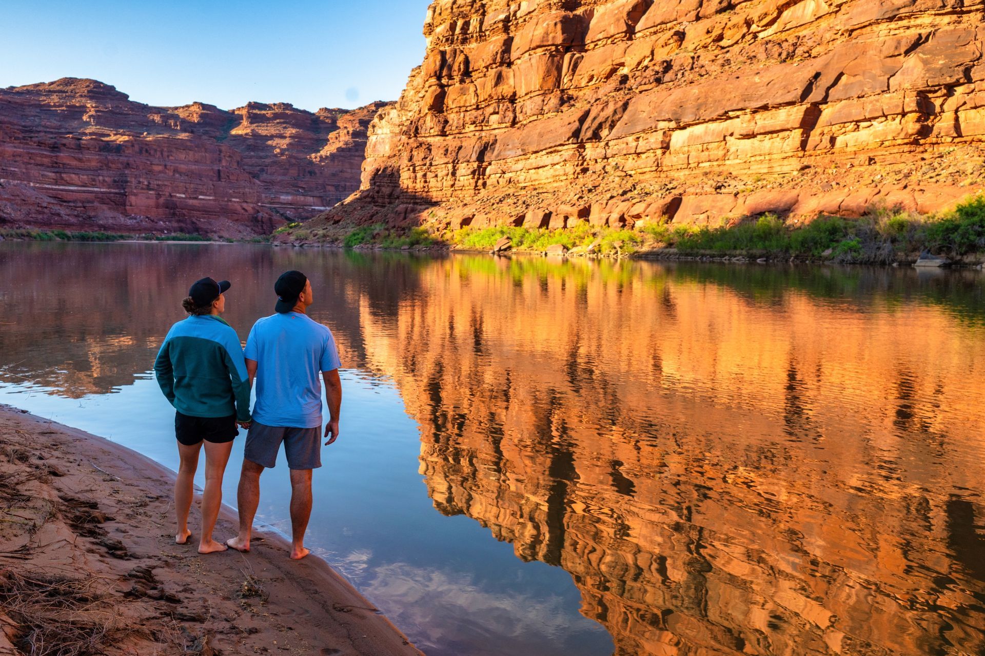 A couple stands beside the Colorado River below sunlit canyon walls