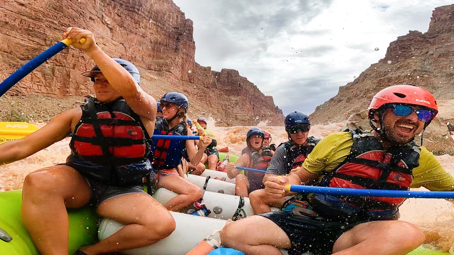 People rafting through Cataract Canyon, wearing helmets and life vests, smiling and paddling on rough water.