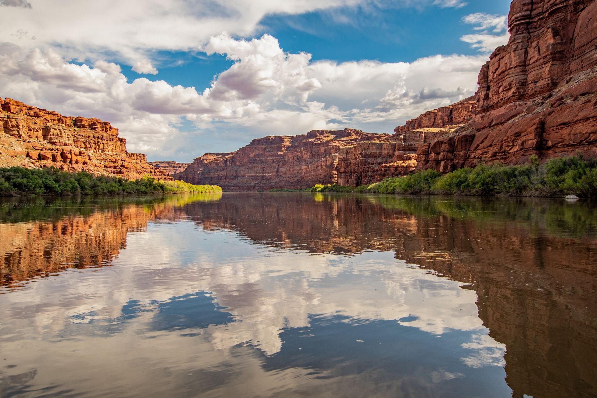 White clouds and blue sky reflect in the water of the Colorado River with green vegetation and towering cliffs on either side.