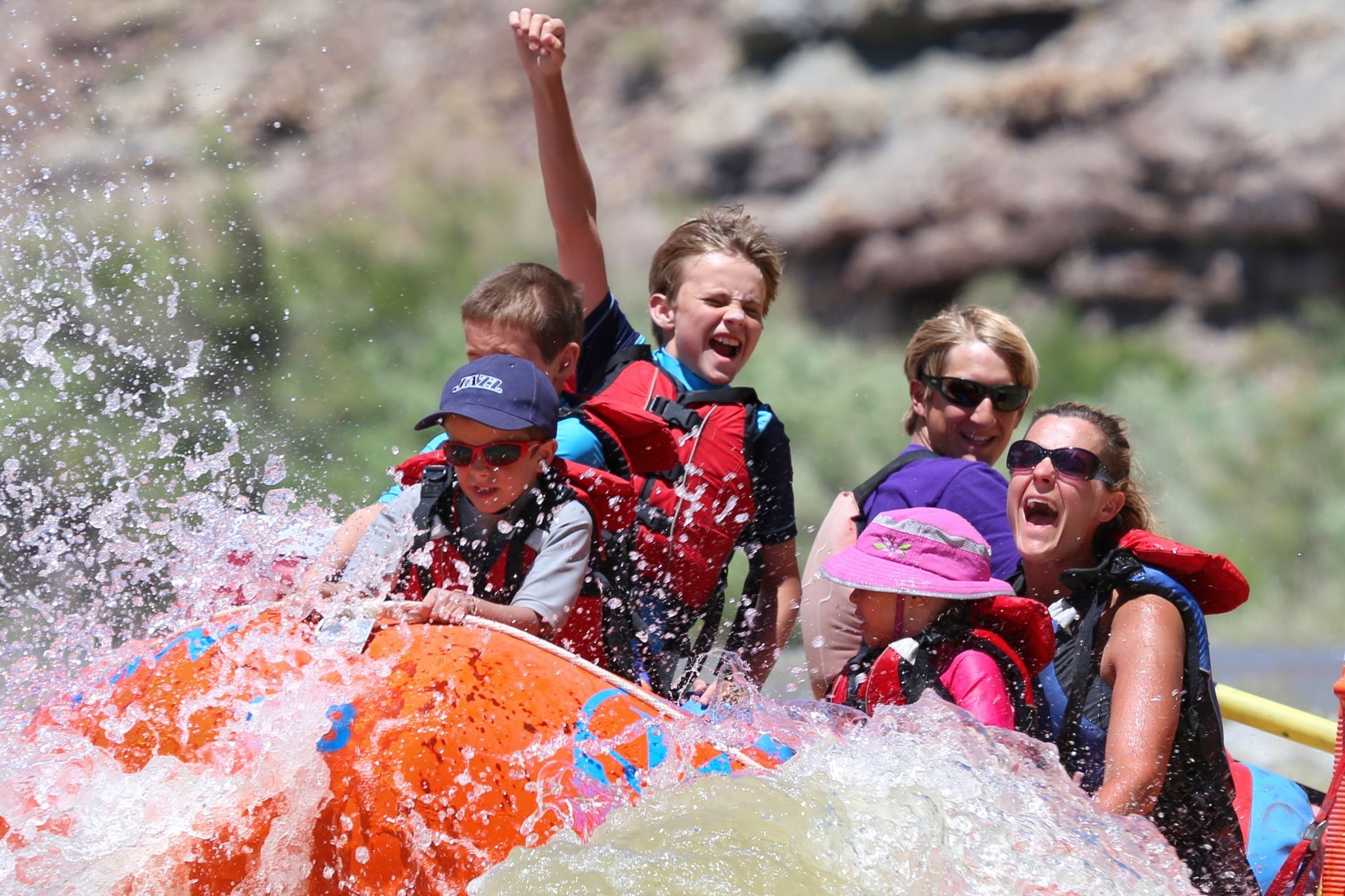 Guests cheer aboard a red oar raft as water splashes around them