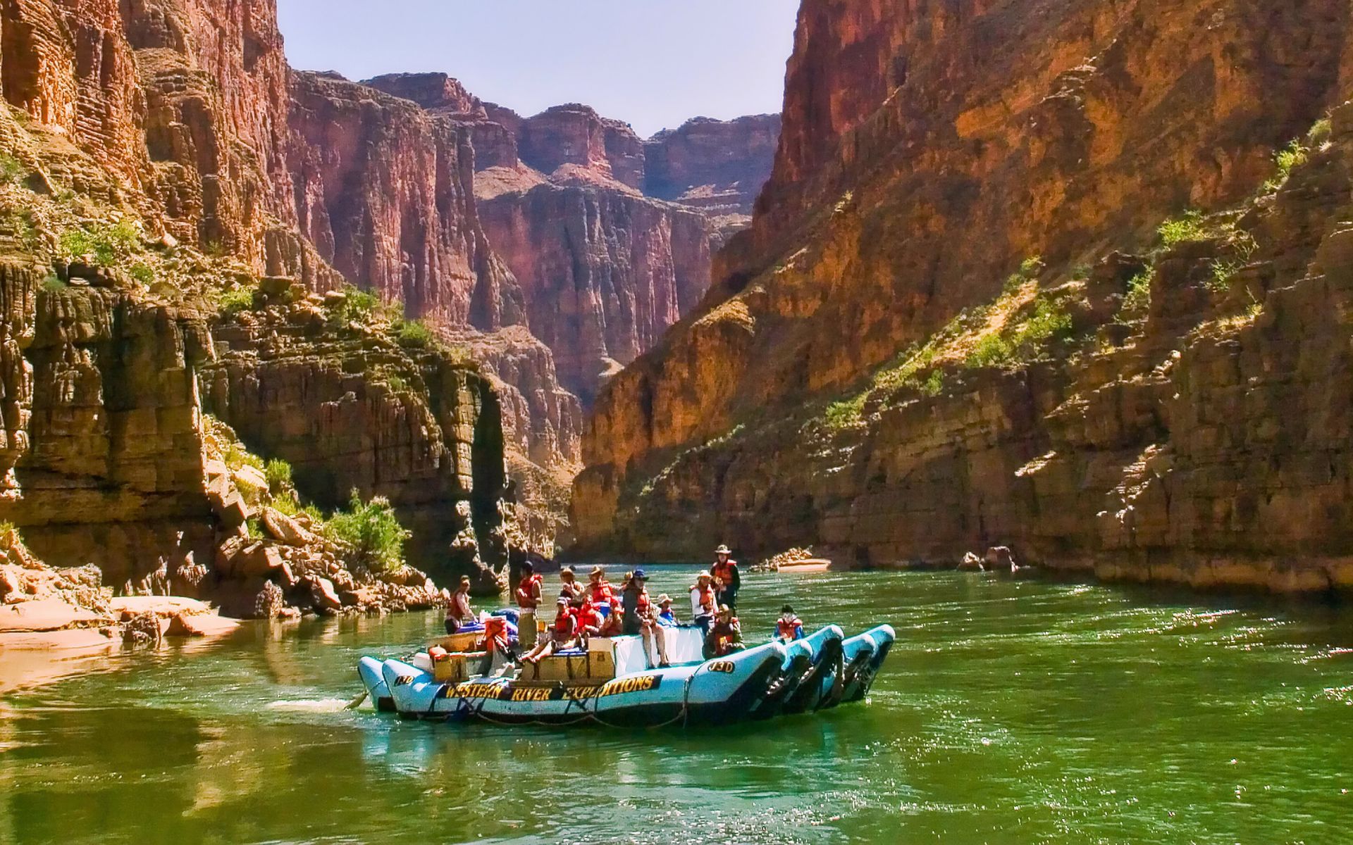 A J-Rig raft floats on emerald green water on the Colorado River with towering cliffs on either side.