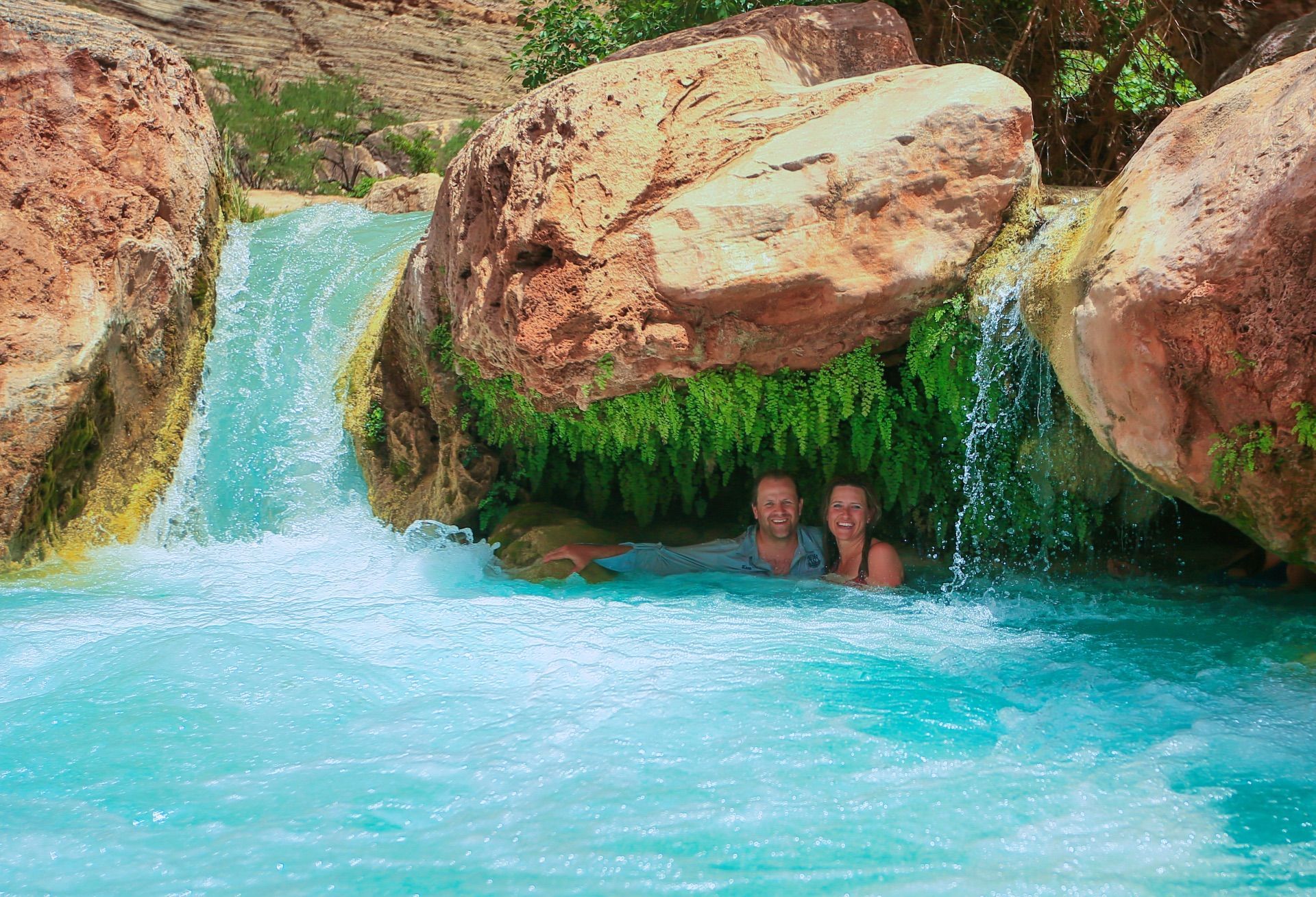Two swimmers beneath a rock ledge beside a Havasu Creek waterfall