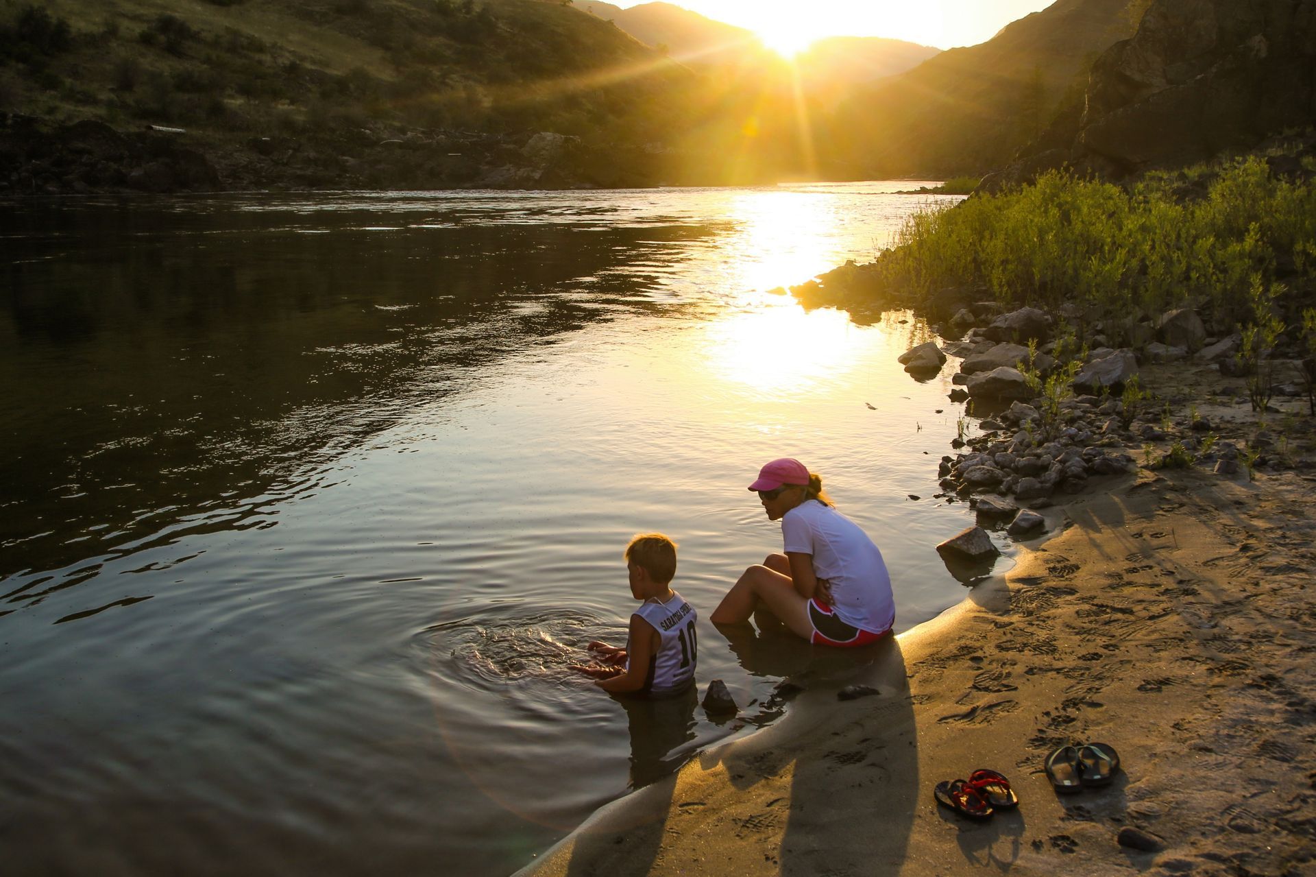 An adult and child sit beside the river at sunset