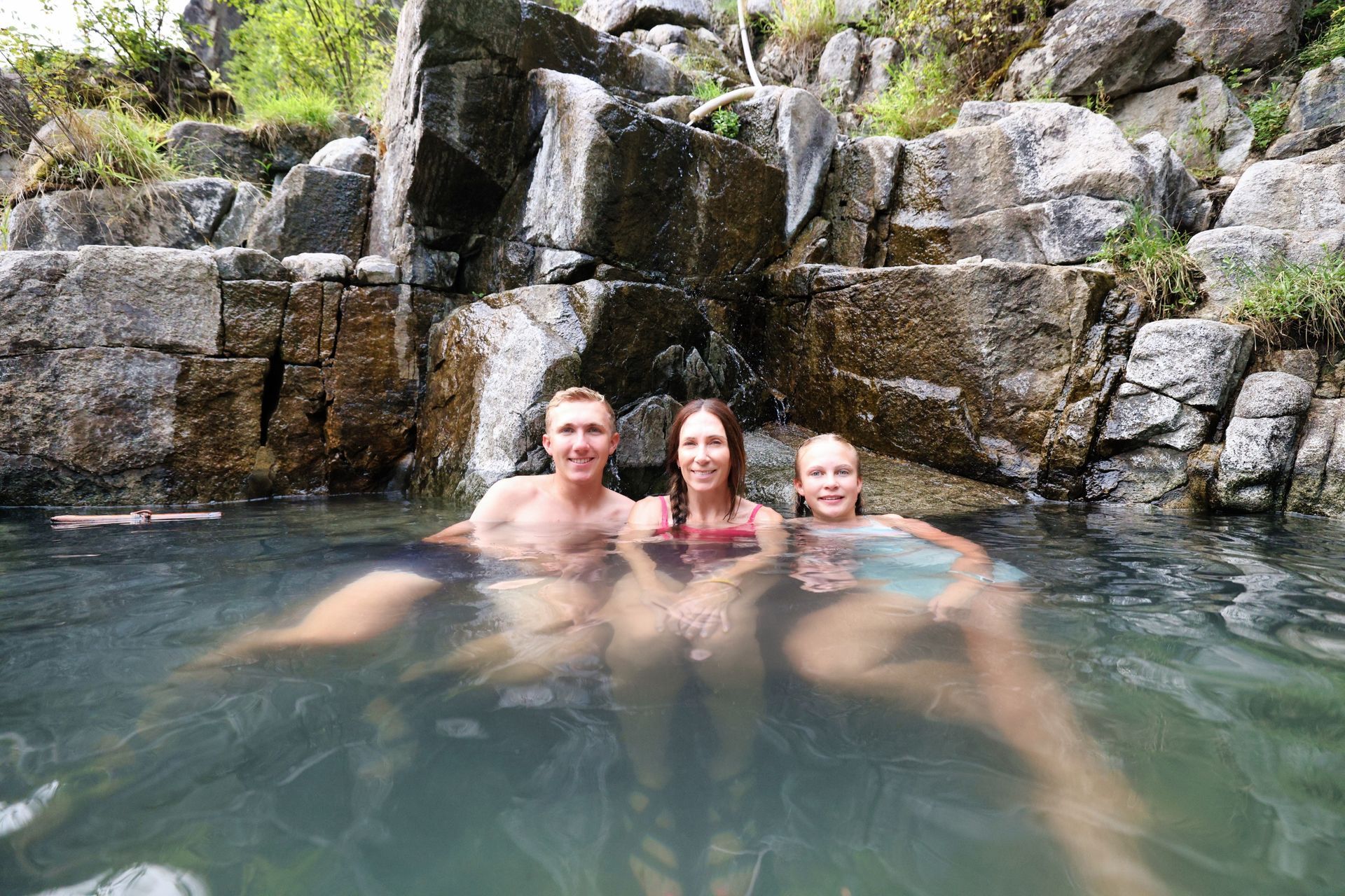 A mother and two children sit in a hot spring pool with a small waterfall behind them.