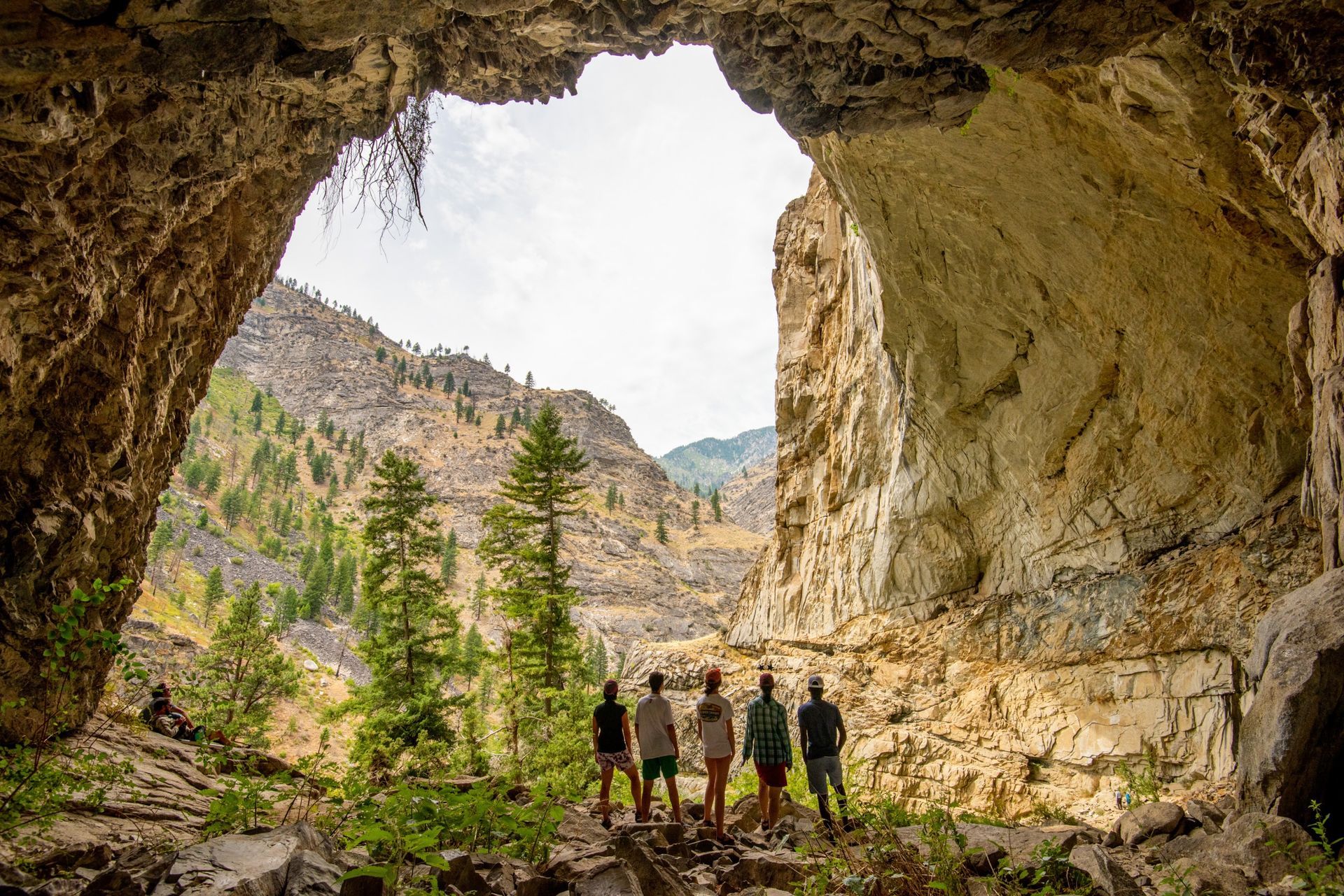 Hikers stand inside a broad rock cave above a forested canyon