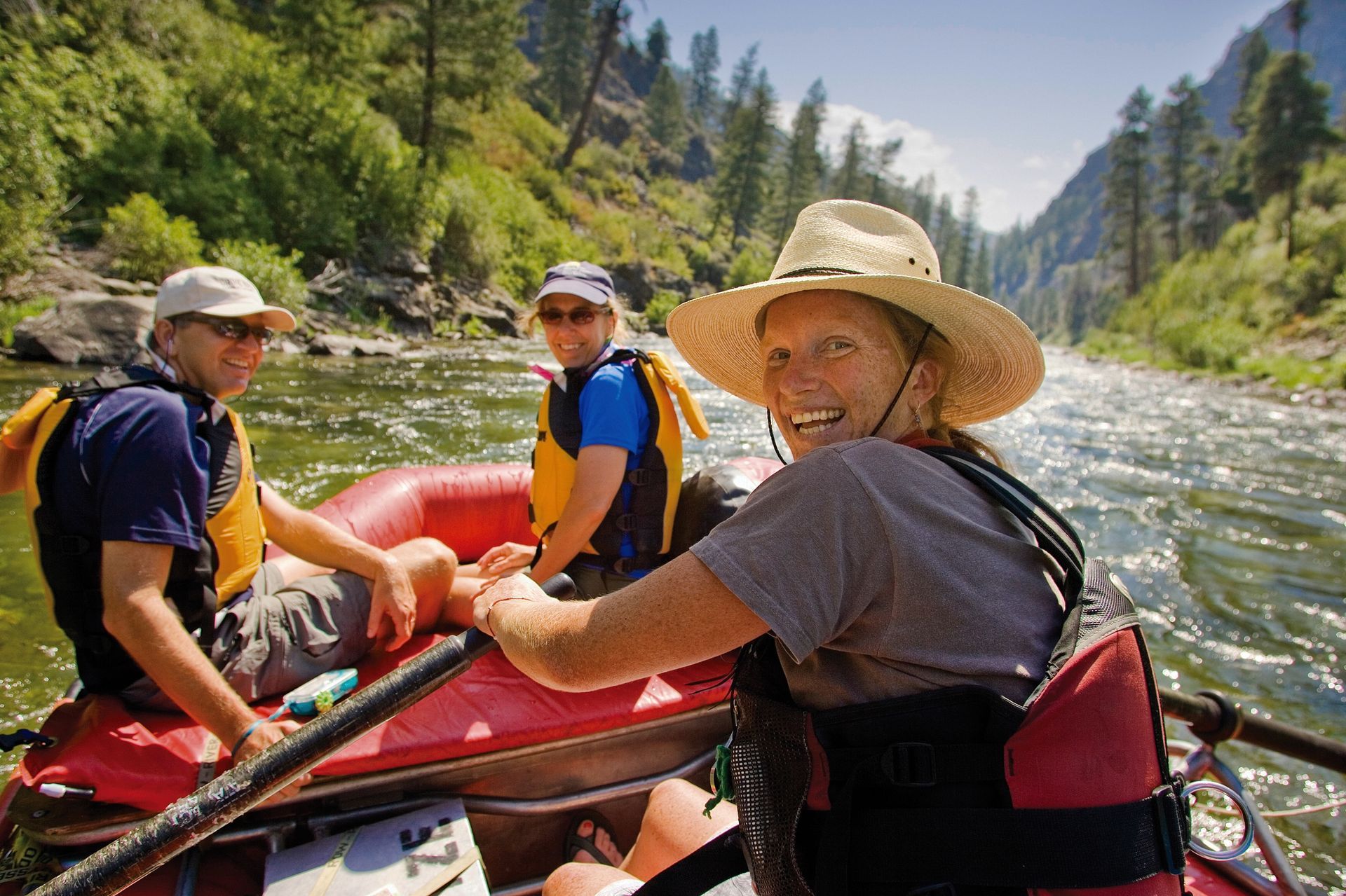 A river guide looks over her shoulder, smiling as she rows a raft, while a couple at the front of the raft smiles at her, with green water and pine trees of an Idaho river in the background.