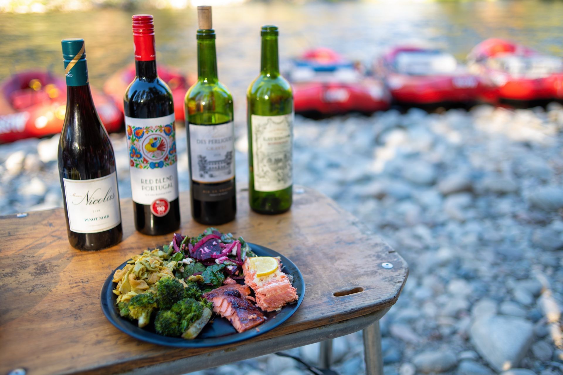 A plate of Salmon, broccoli, pasta and salad on a table with 4 wine bottles and a rocky beach with rafts at the rivers edge in the background
