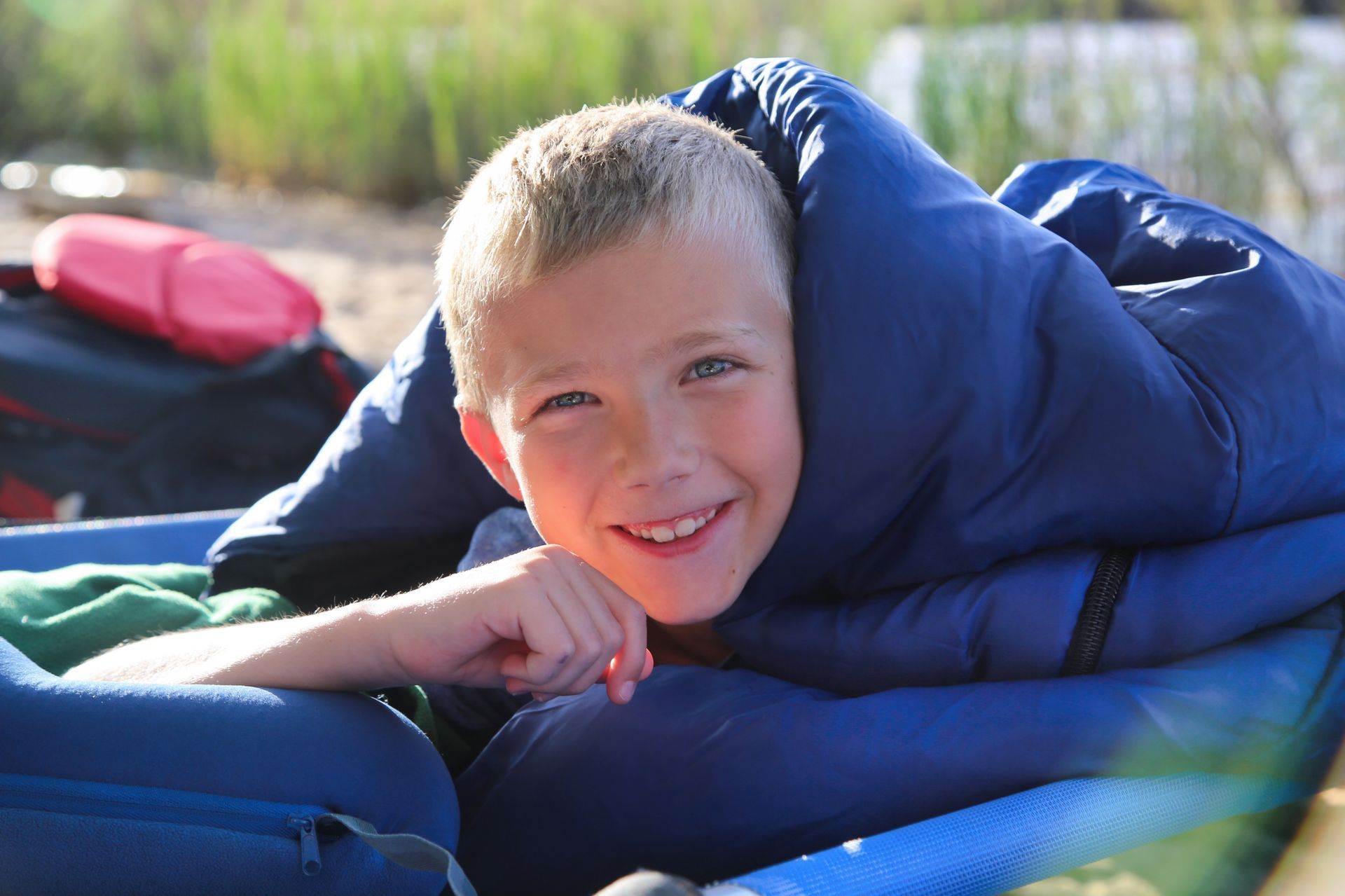 A boy looks up from his sleeping bag while laying on a cot at the river's edge.