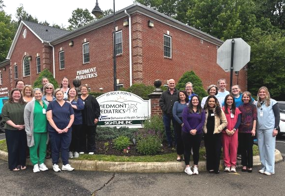 Group of people posing outside a brick building beside a sign, with trees and a cloudy sky behind them.