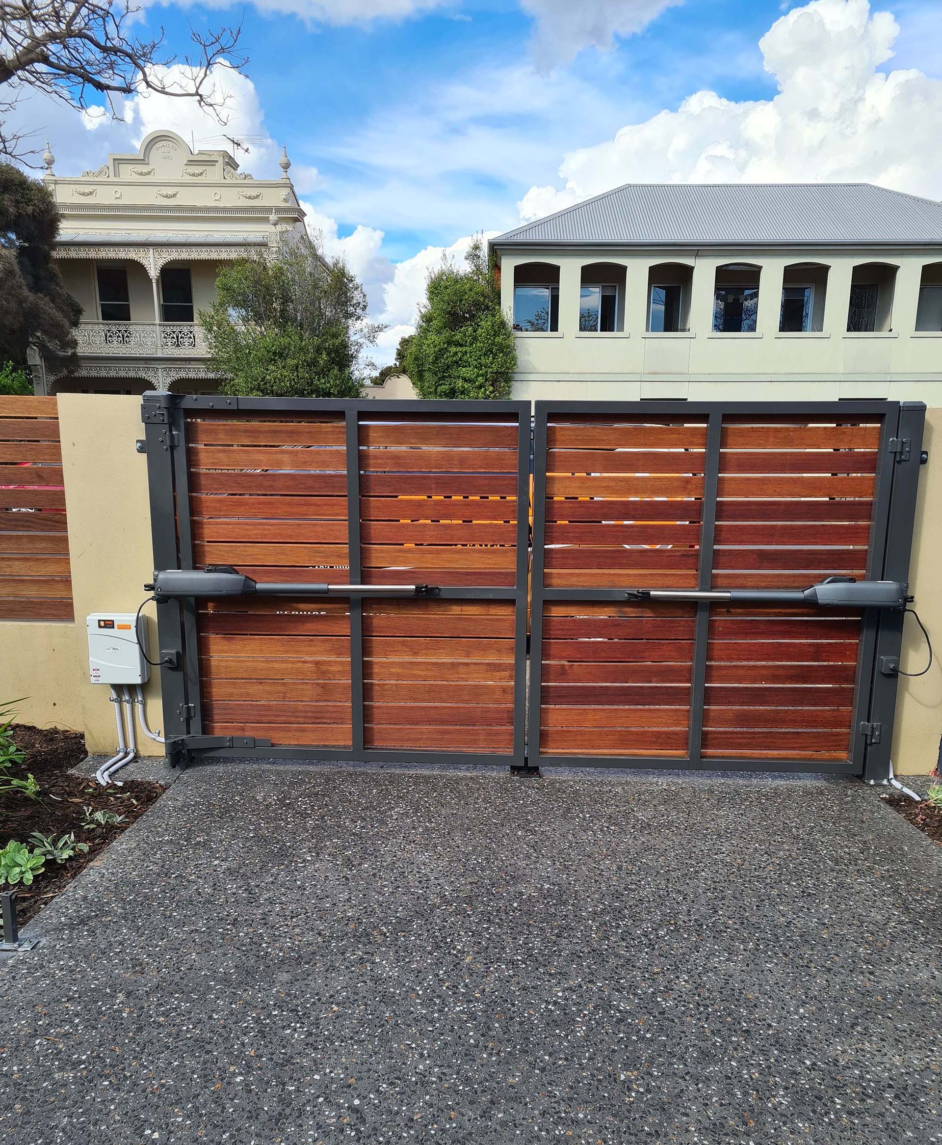 Black metal pedestrian gate with pointed bars beside a textured wall and driveway.