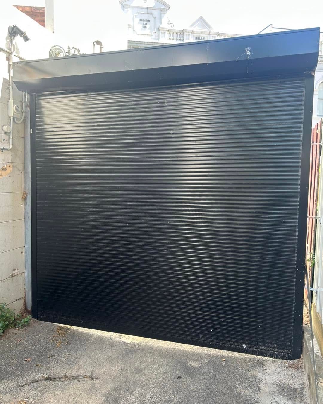 Black corrugated metal roll-up gate on a narrow driveway or alley entrance