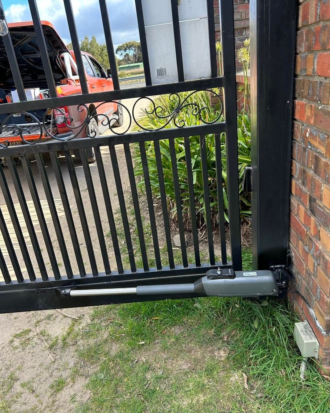 Black metal gate with a silver automatic opener beside a brick pillar and driveway.
