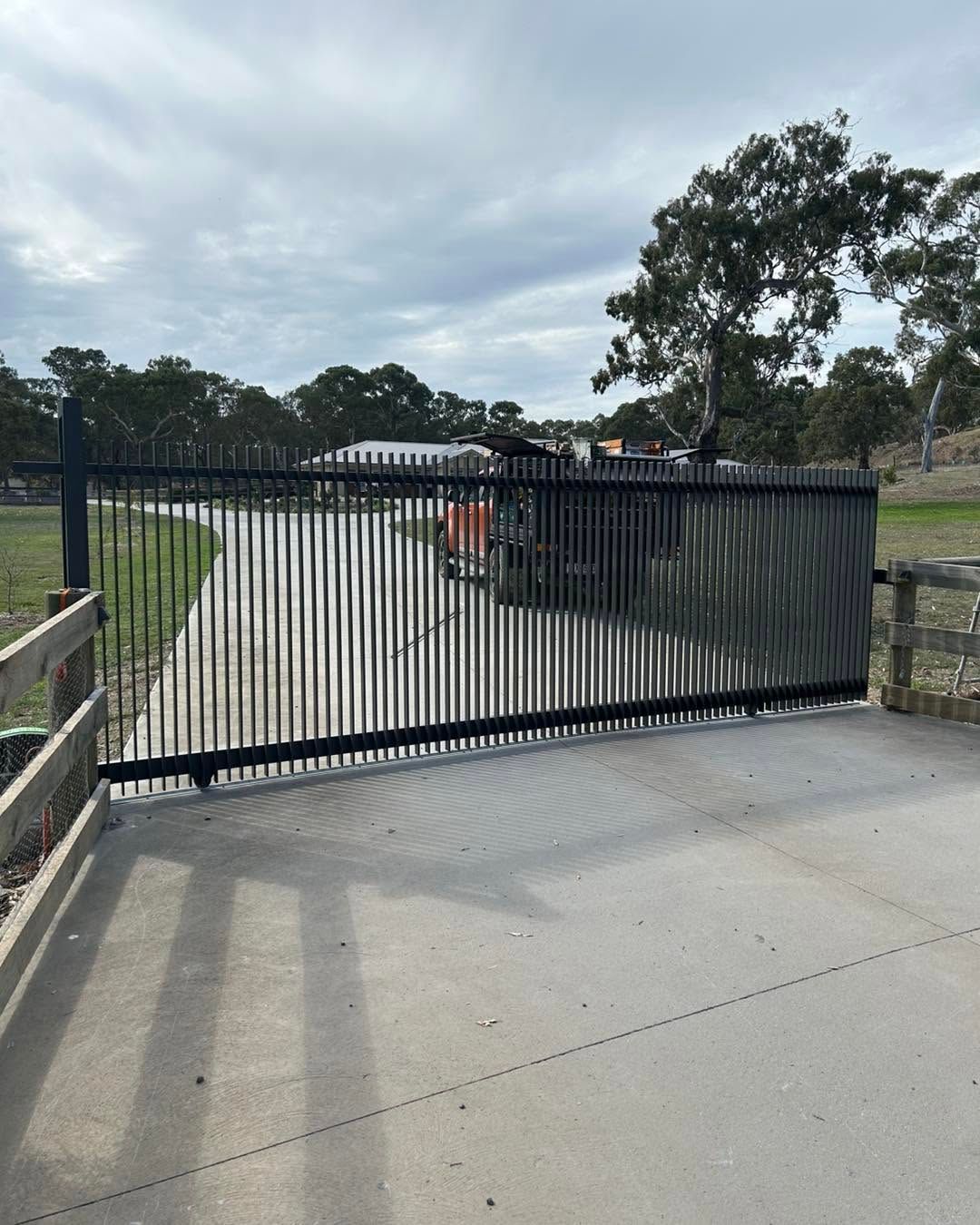 Closed black metal gate blocking a concrete driveway, with trees and a cloudy sky in the background