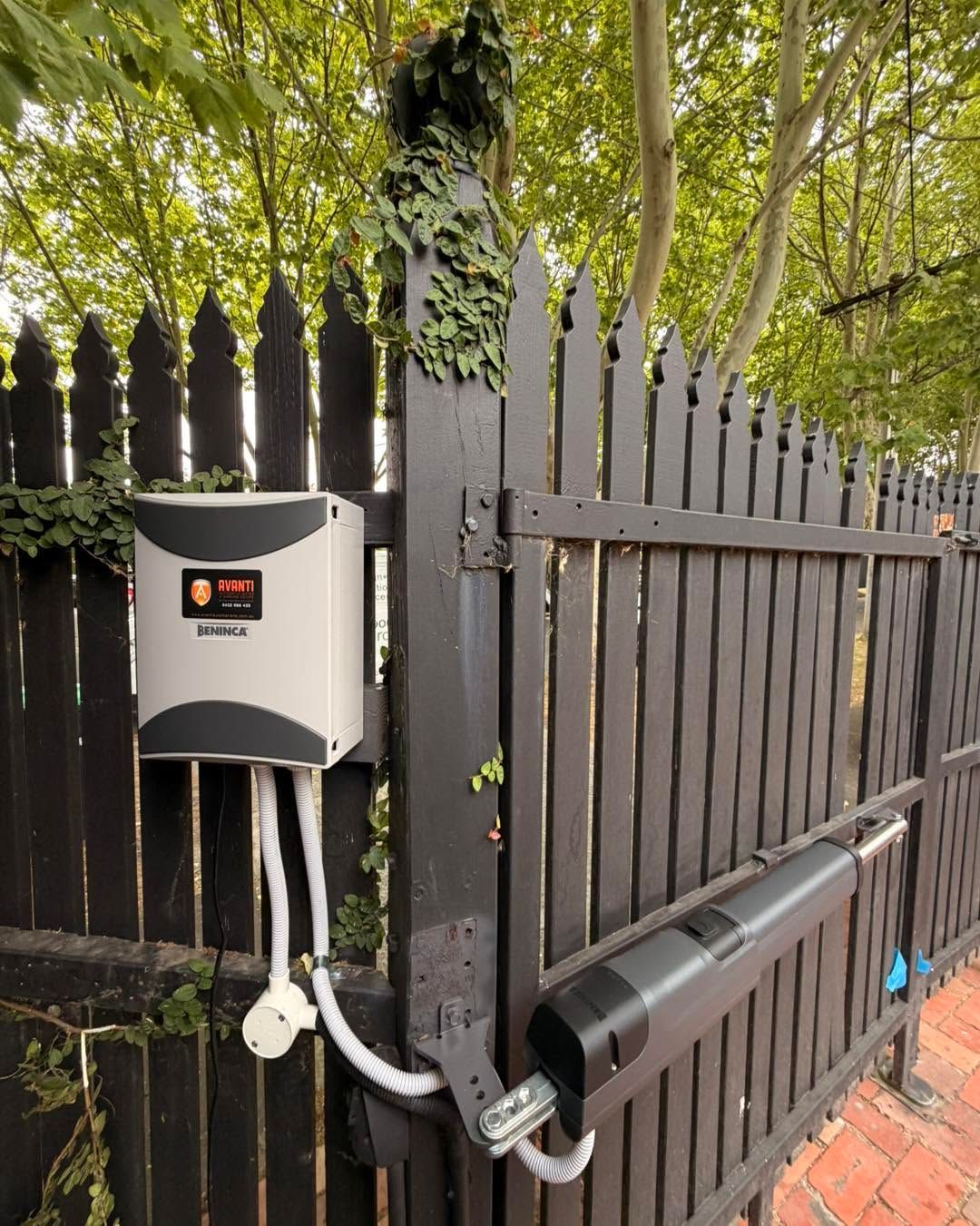Black metal fence with a wall-mounted control box and conduit beside a brick walkway under trees