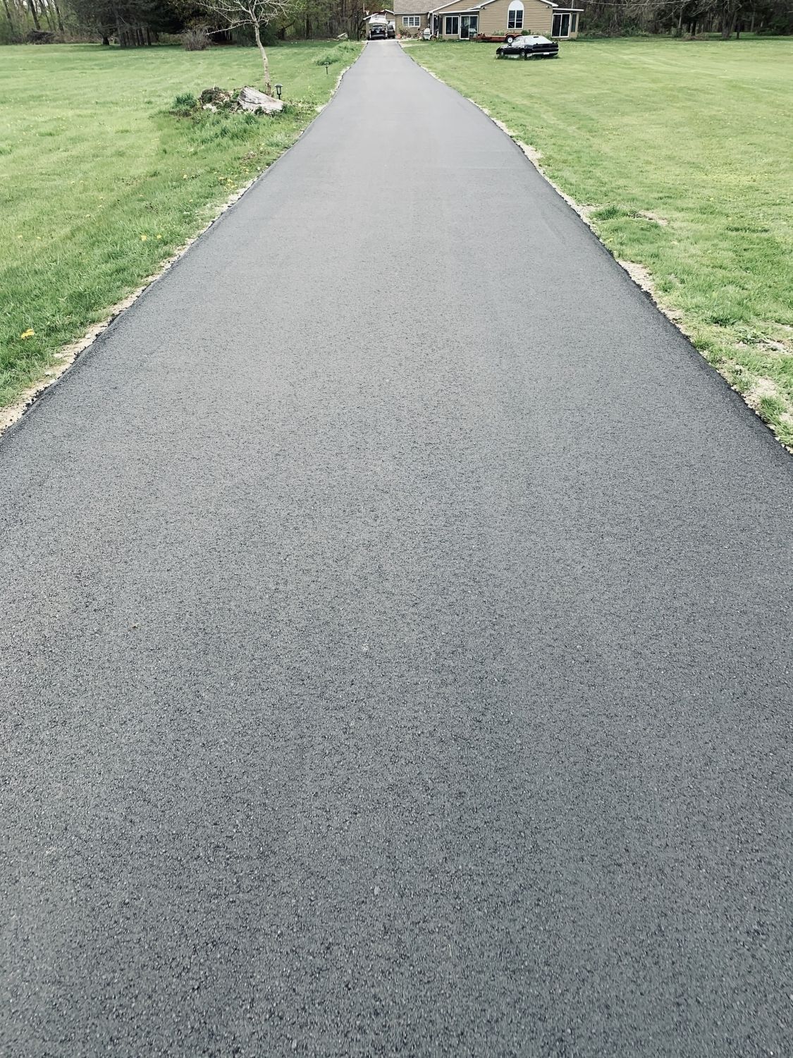 A long asphalt driveway leading to a house in a grassy field.