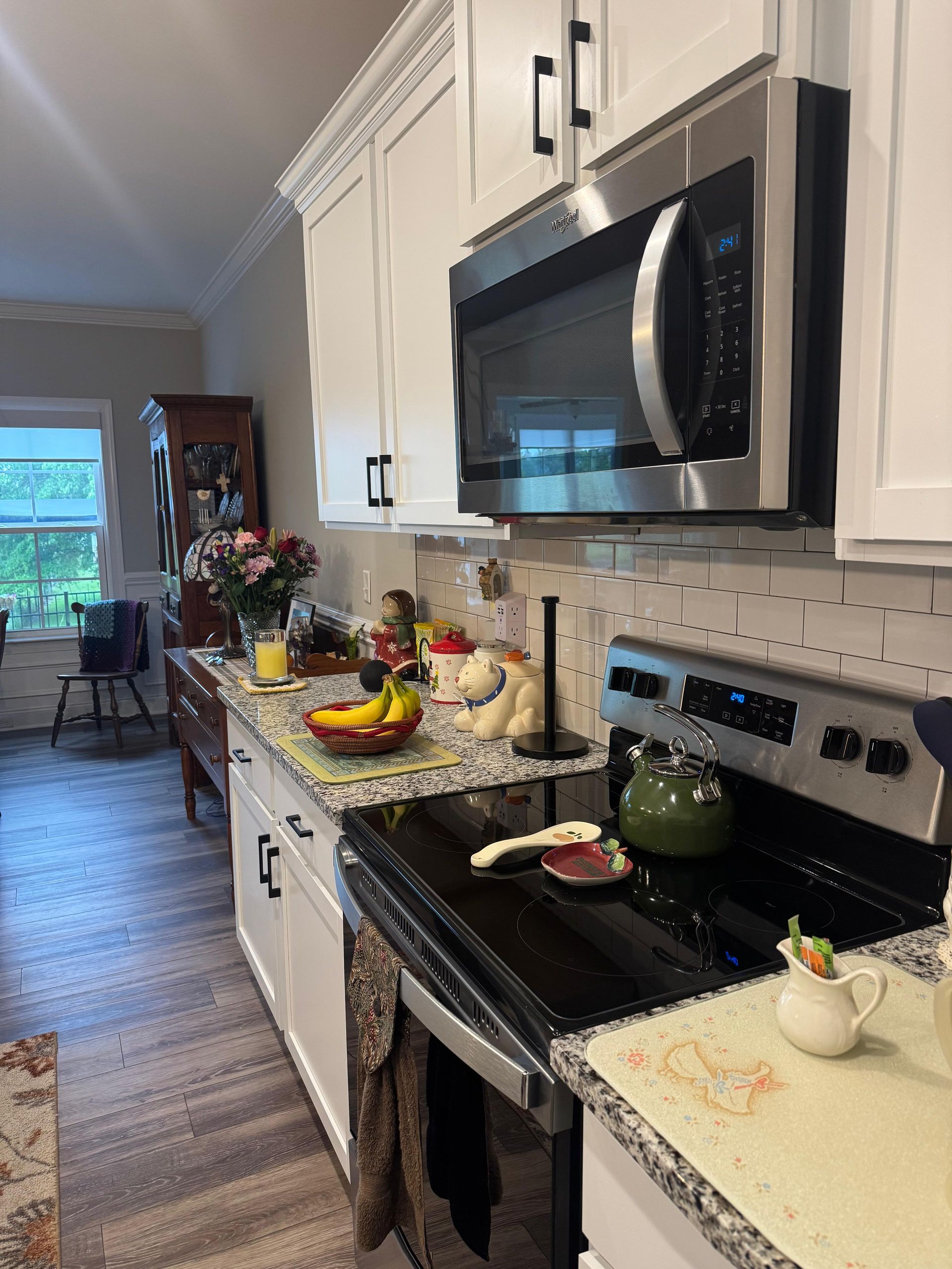 Bright kitchen with white cabinets, black stove and microwave, and fruit on a granite counter leading to a dining area.
