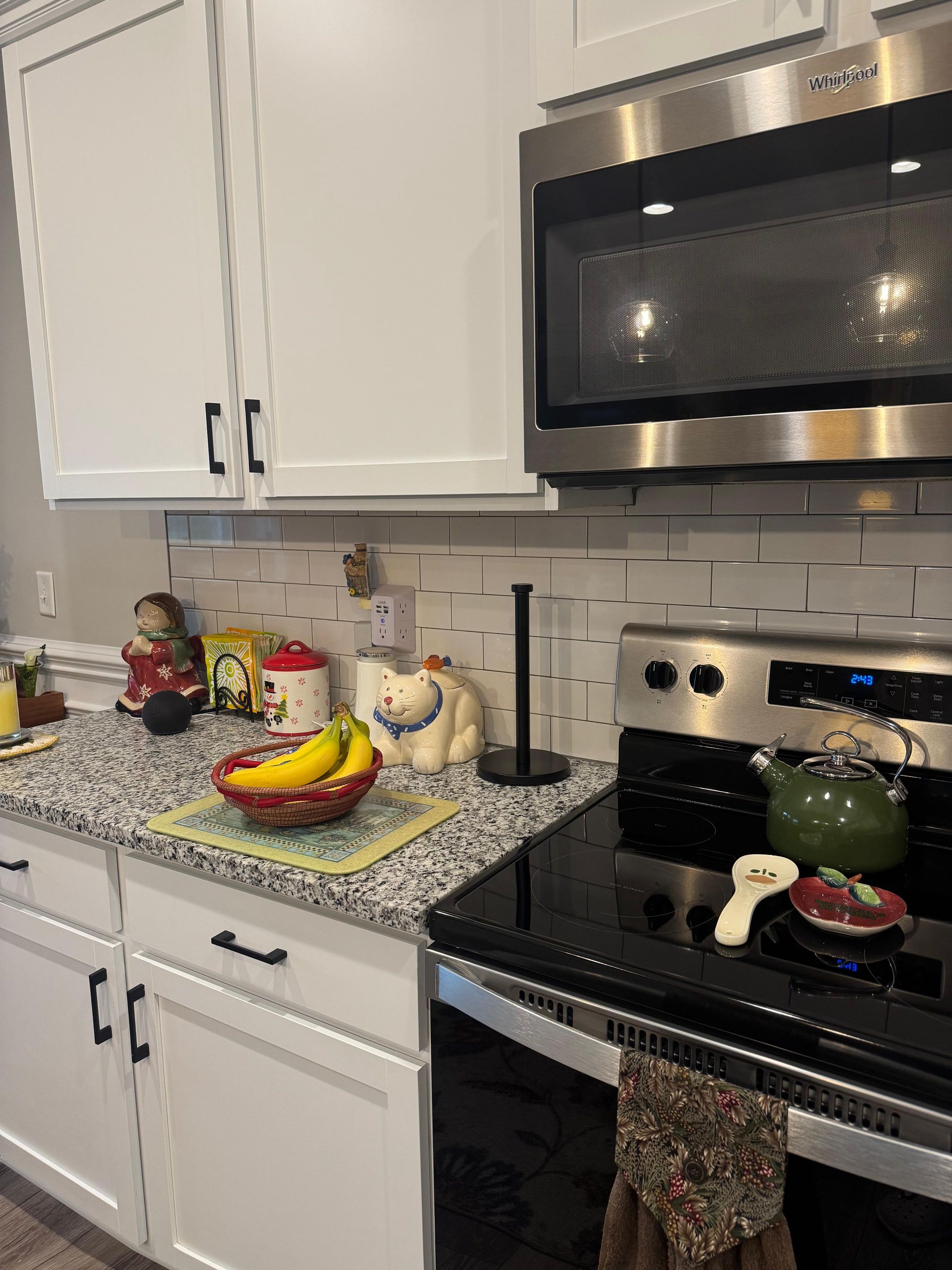 Kitchen counter with fruit, jars, a kettle, and a green pot on a stove under white cabinets and a microwave. Kitchen counter with fruit, jars, a kettle, and a green pot on a stove under white cabinets and a microwave.