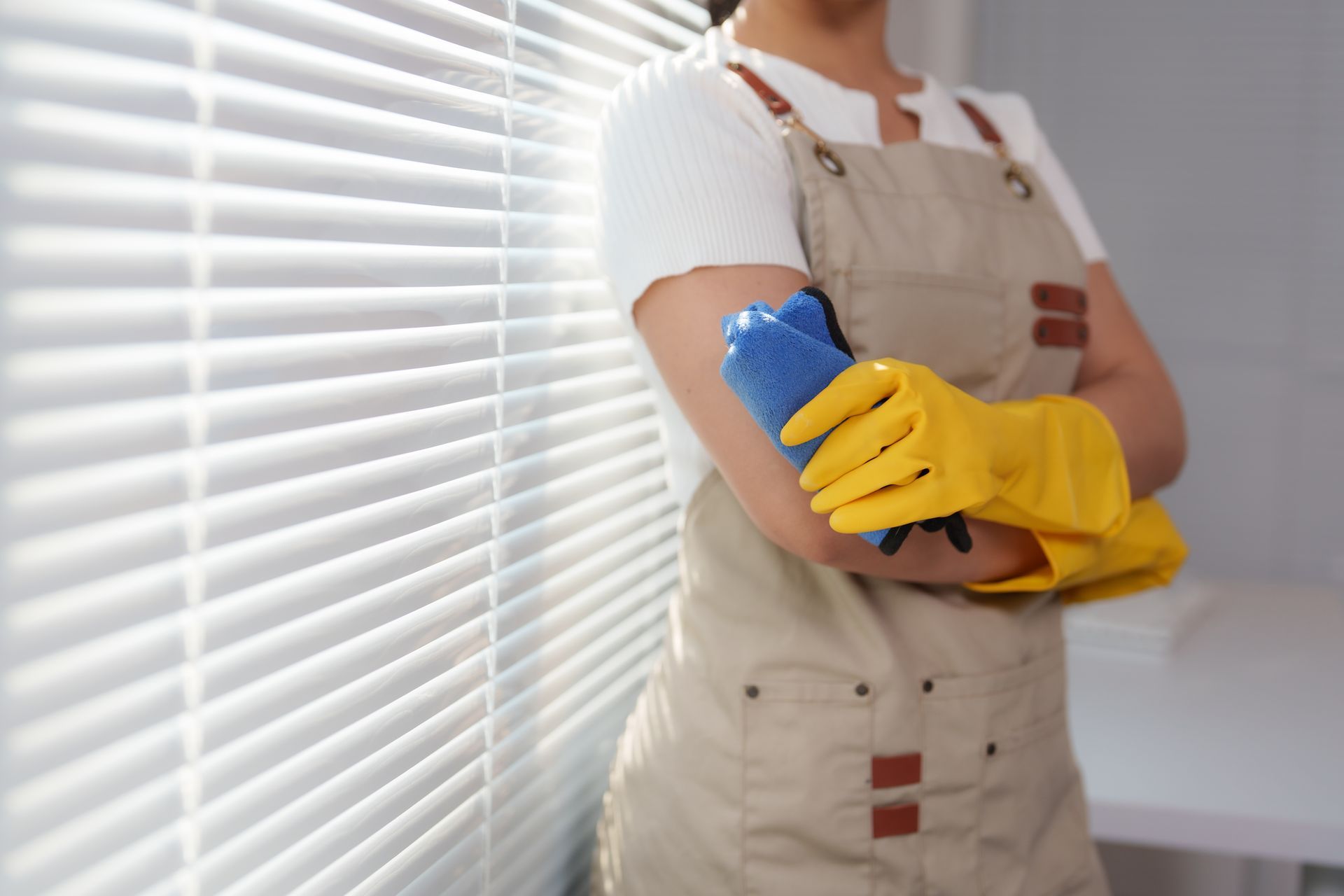Person in apron and yellow gloves cleaning a window blind with a blue cloth. Person in apron and yellow gloves cleaning a window blind with a blue cloth.