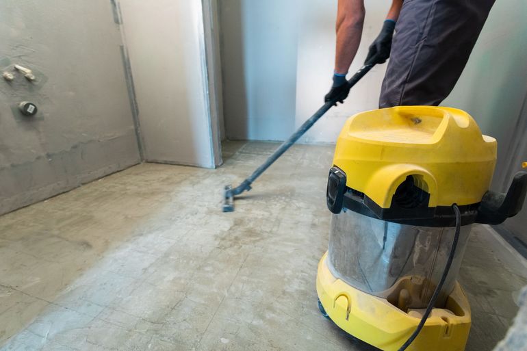 Person vacuuming a dusty concrete floor with a yellow wet-dry vacuum in an unfinished room.