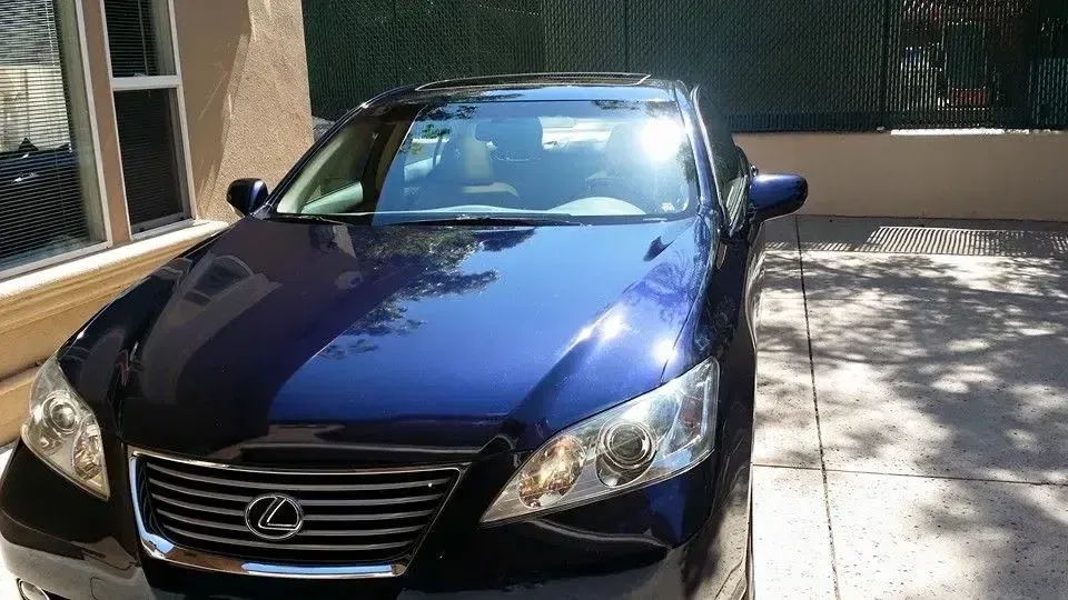 Blue Lexus sedan parked in a driveway beside a building and fence, with sunlight and shadows.