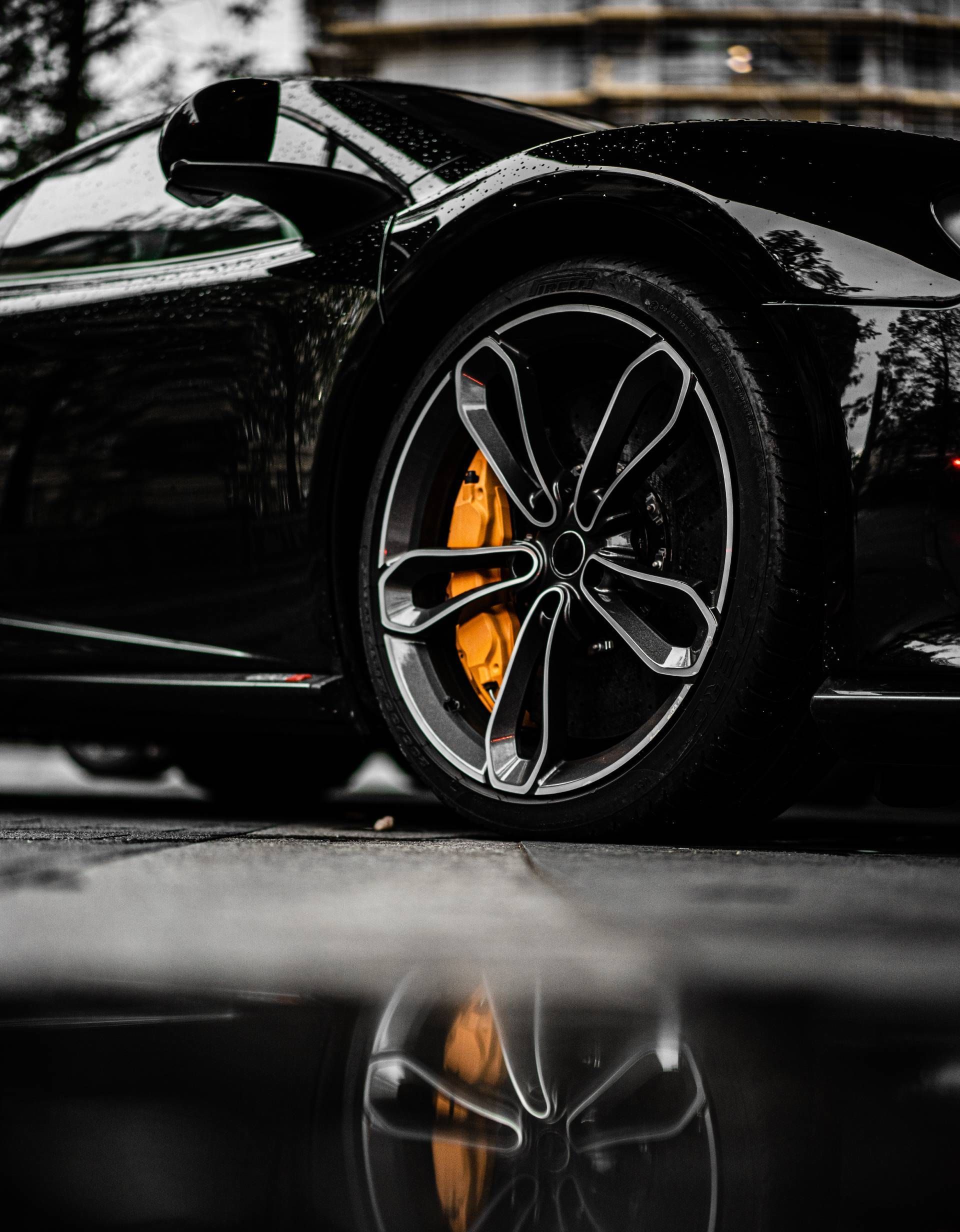 Close-up of a black sports car wheel with orange brake caliper reflected on a wet surface