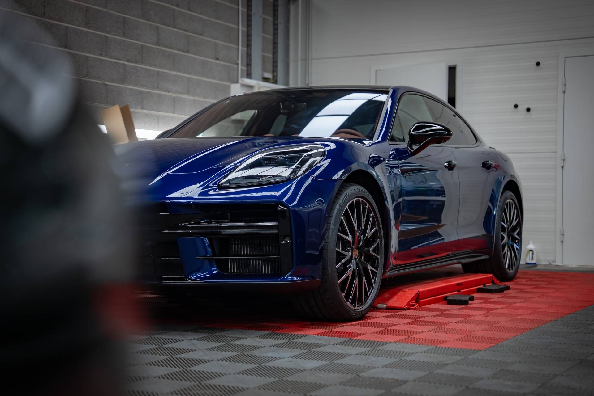 Blue sports sedan on a red platform in a garage, with bright headlights and a glossy finish.