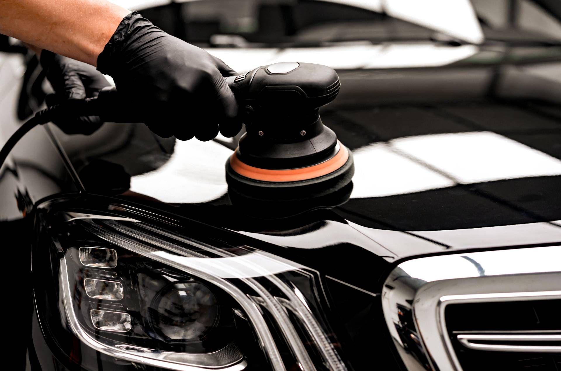 Gloved hand polishing a black car hood with a buffer, close-up of the front headlight and grille.
