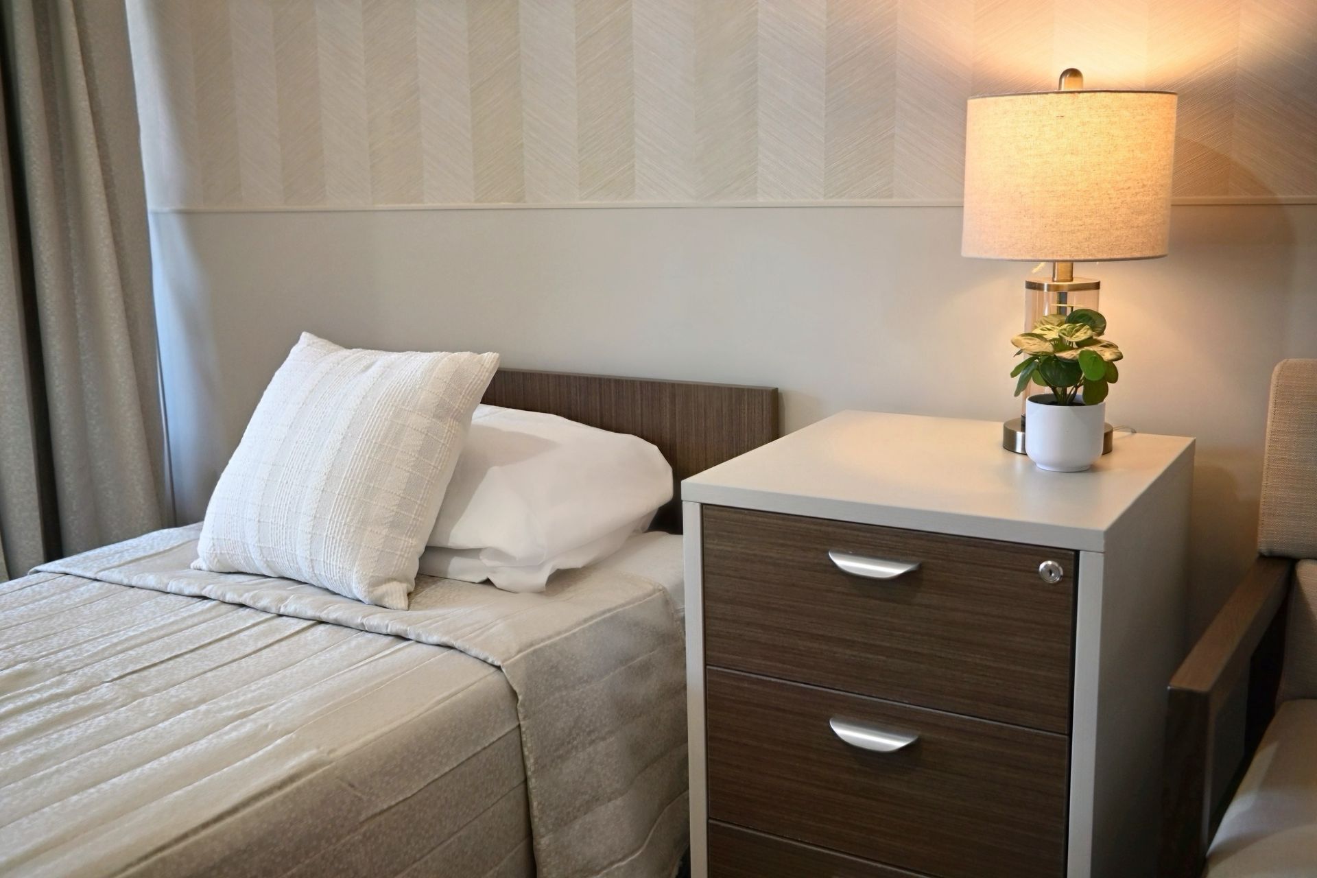Neatly made bed beside a white nightstand with lamp and potted plant in a small bedroom