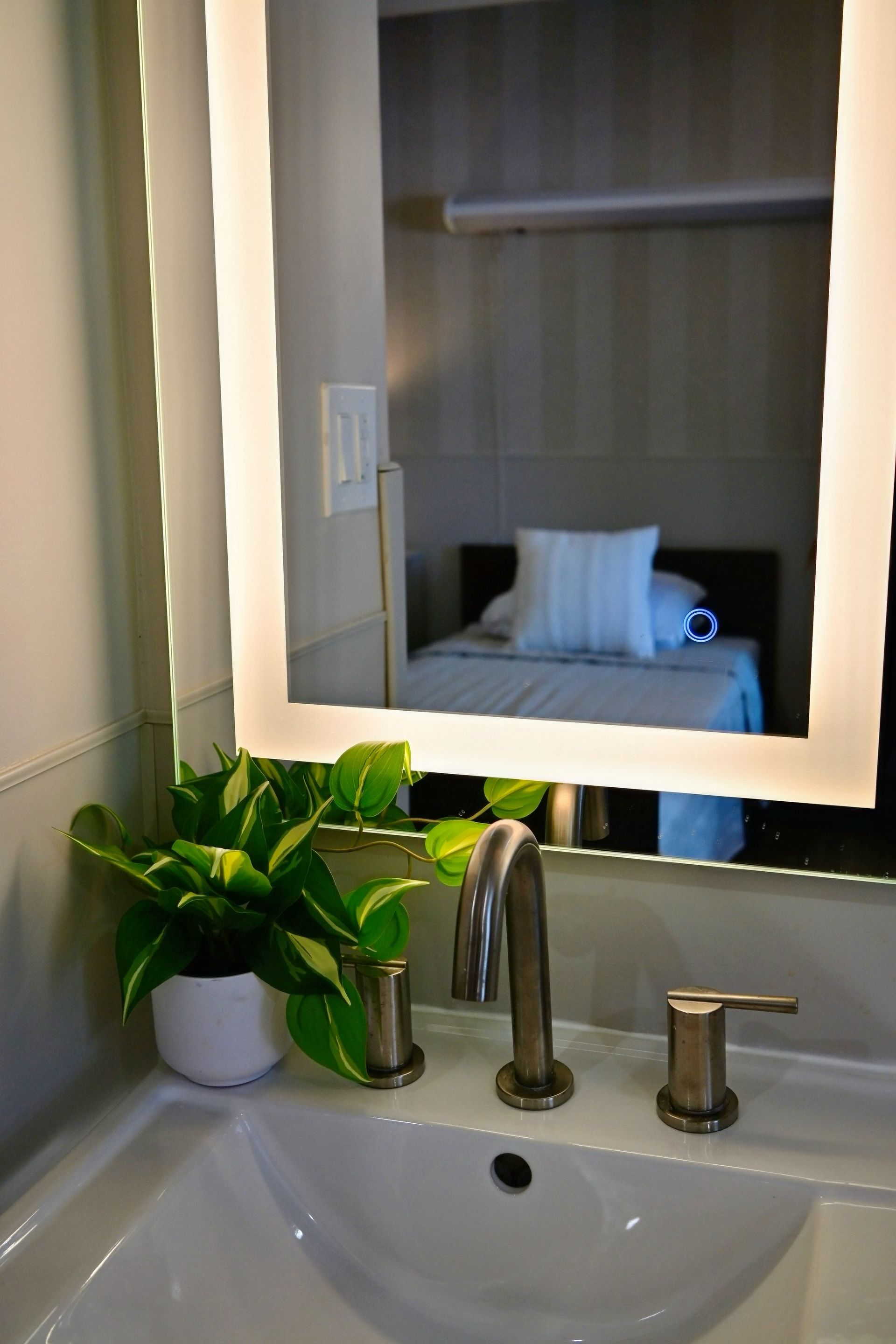 Bathroom sink with lit mirror reflecting a bed in a bedroom beyond, and a potted plant on the counter