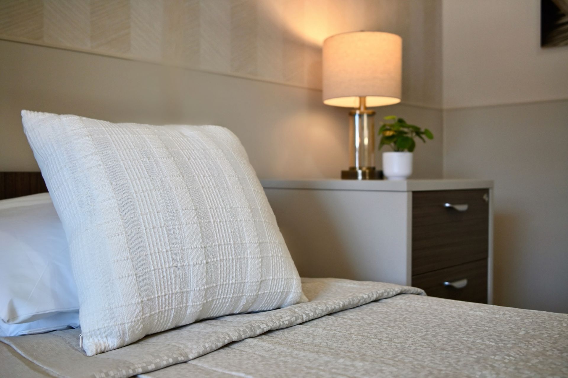 White pillow on neatly made bed beside a nightstand with lamp in a softly lit bedroom