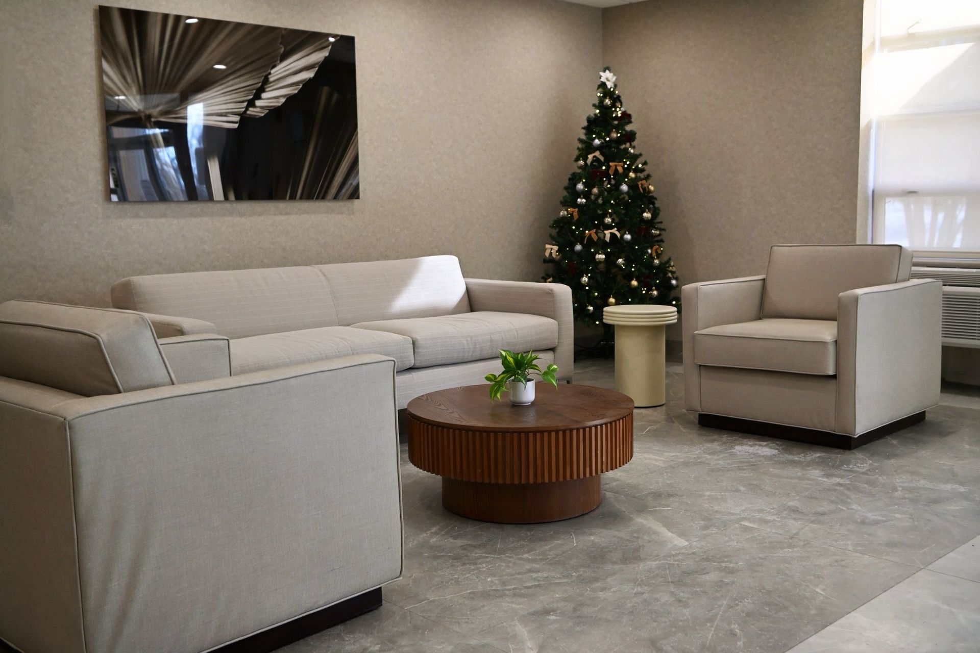 Modern living room with beige sofa and chairs, round wooden table, gray rug, and a decorated Christmas tree.