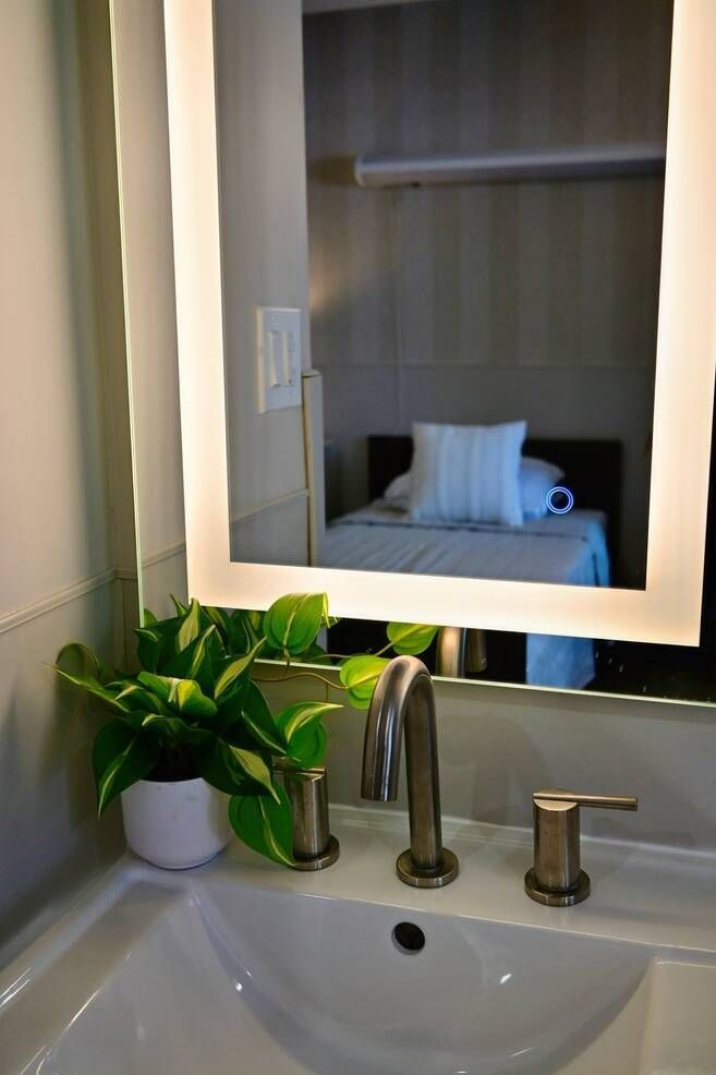 Bathroom sink with lit mirror reflecting a hotel-style bed and pillow, with a green plant beside the faucet