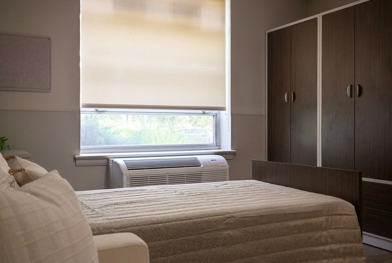 Bedroom with bed, window air conditioner, beige blinds, and dark wood wardrobe
