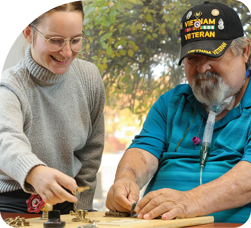 Two people smiling while playing chess at a table indoors; one wears a navy veteran cap.