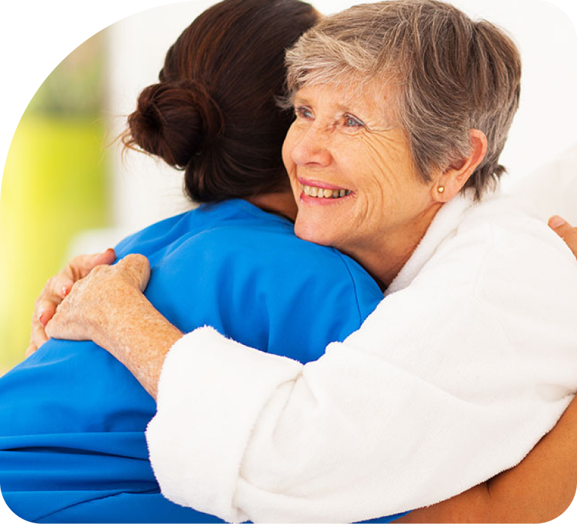 Two people hugging, one in a white sweater and one in blue, smiling warmly indoors