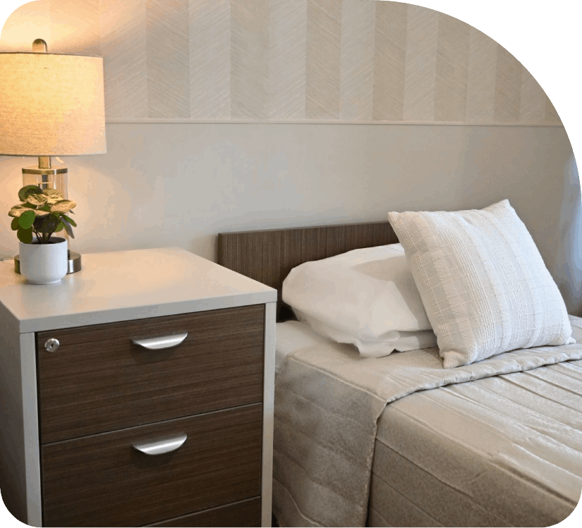 Neatly made bed beside a dark wood nightstand with lamp and plant in a softly lit bedroom