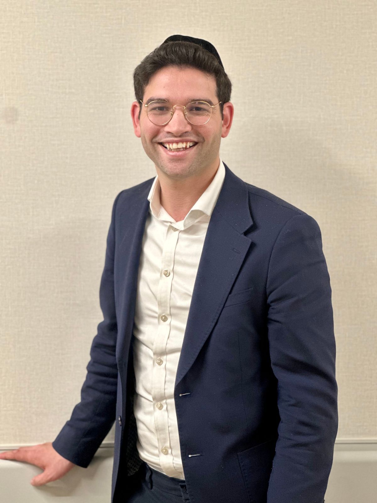 Smiling person in a navy blazer and white shirt standing against a beige wall