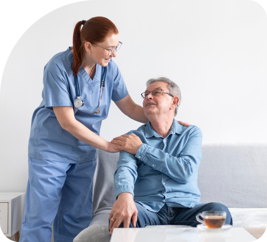 Nurse in blue scrubs helping a seated patient on a couch in a bright living room
