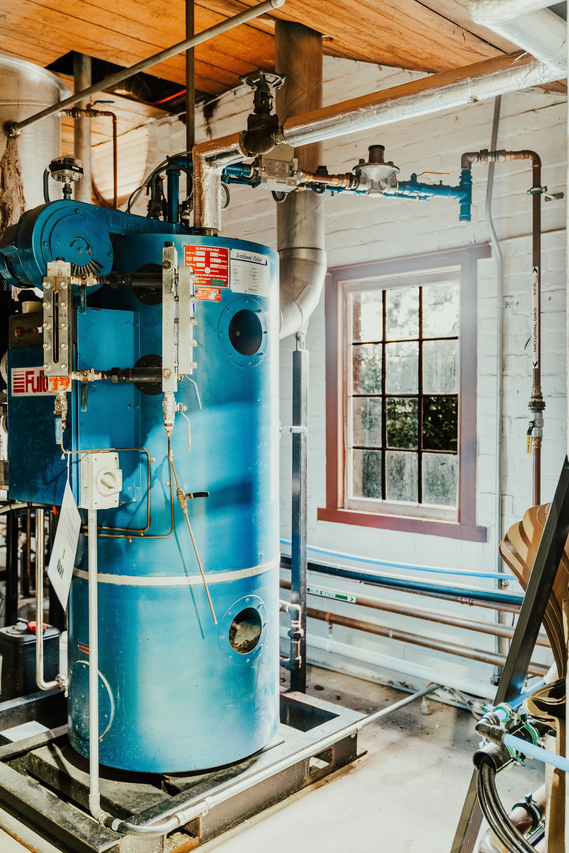 Blue industrial water heater with pipes in a room with a window.