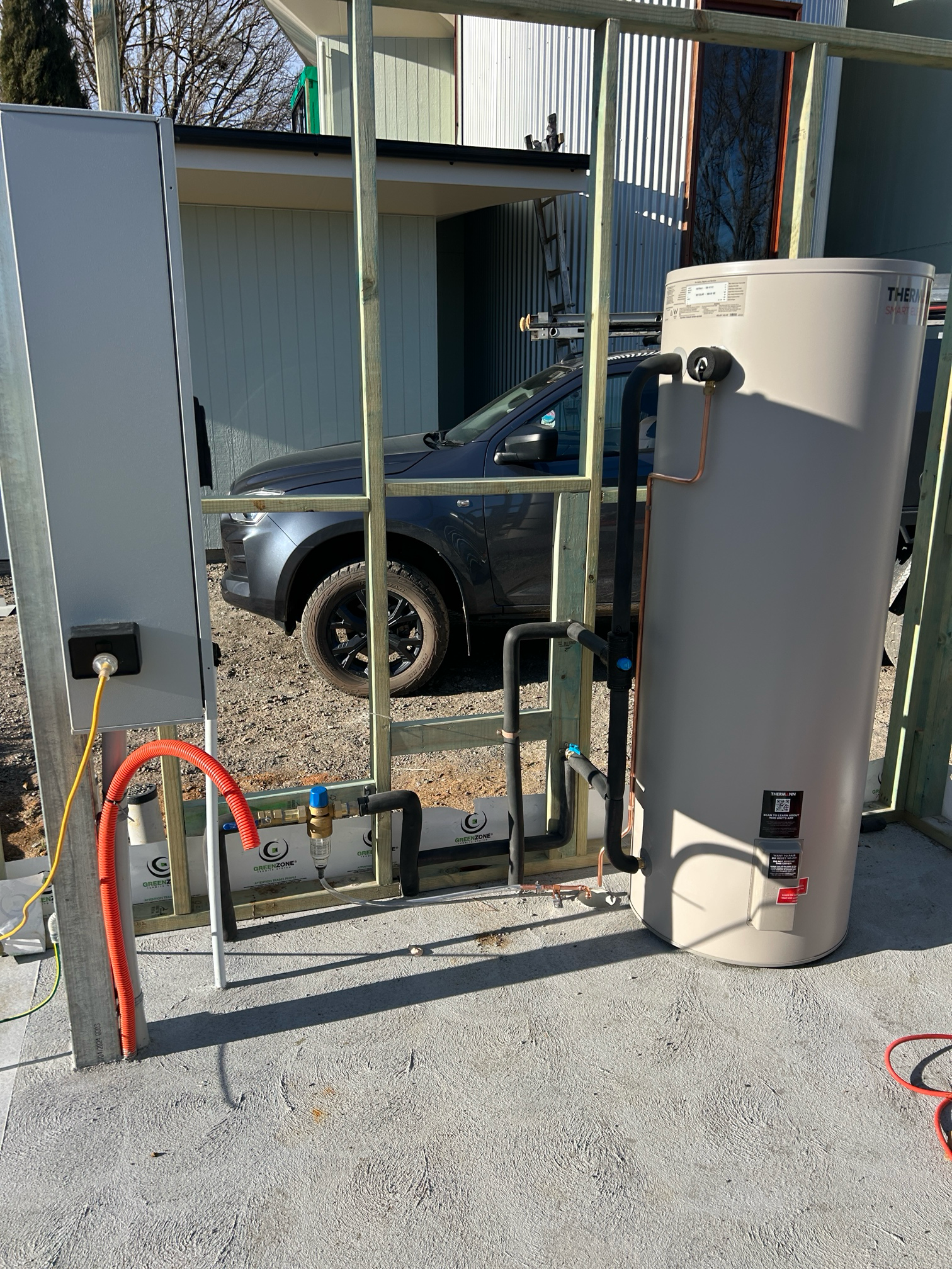 Water heater system with piping and electrical box, outdoor setting. Gray tank, black and red pipes.