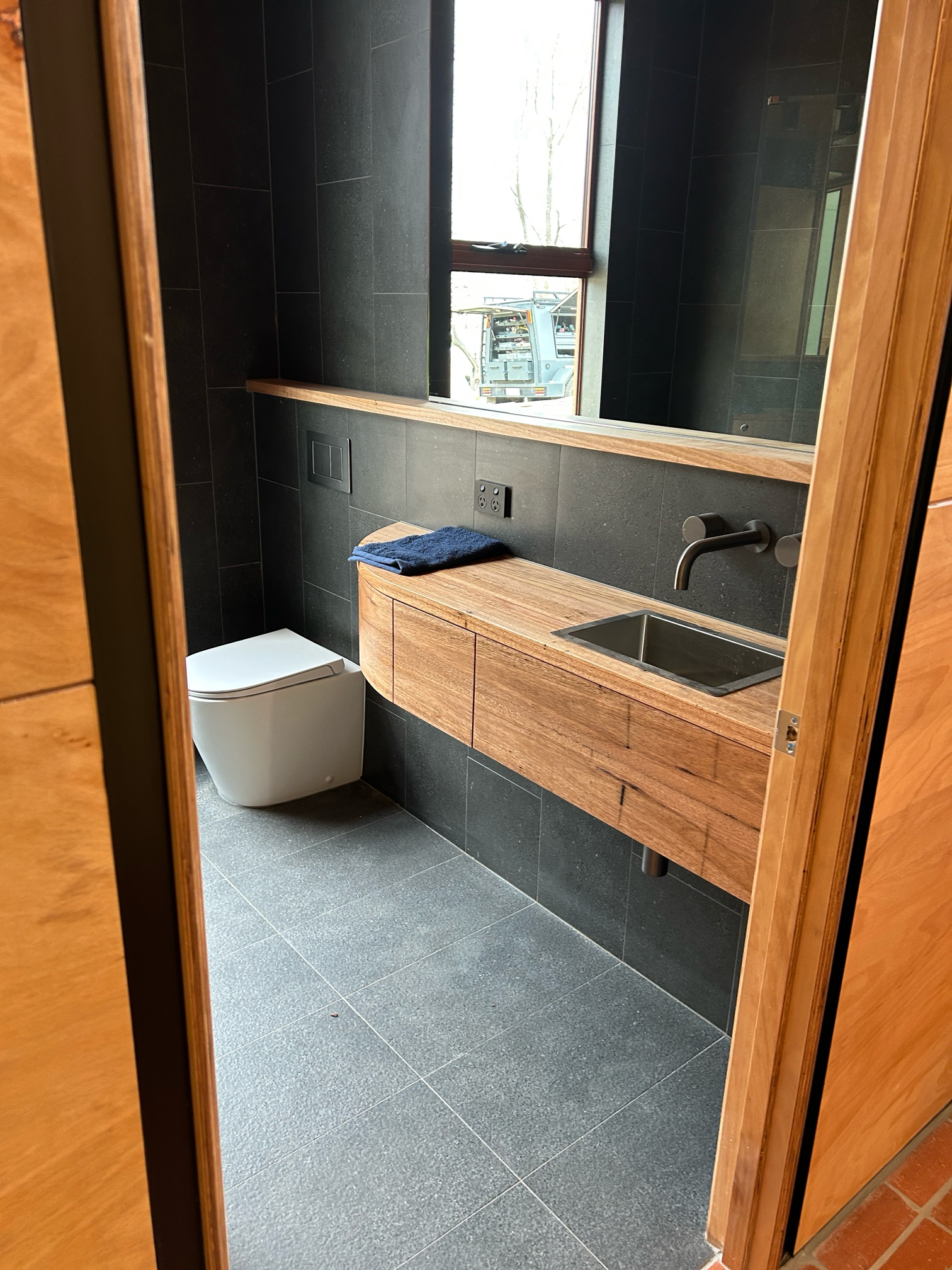 Modern bathroom with black tile walls, wooden vanity, and white toilet.