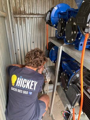 Worker crouches beside a blue industrial machine inside a corrugated metal shed.