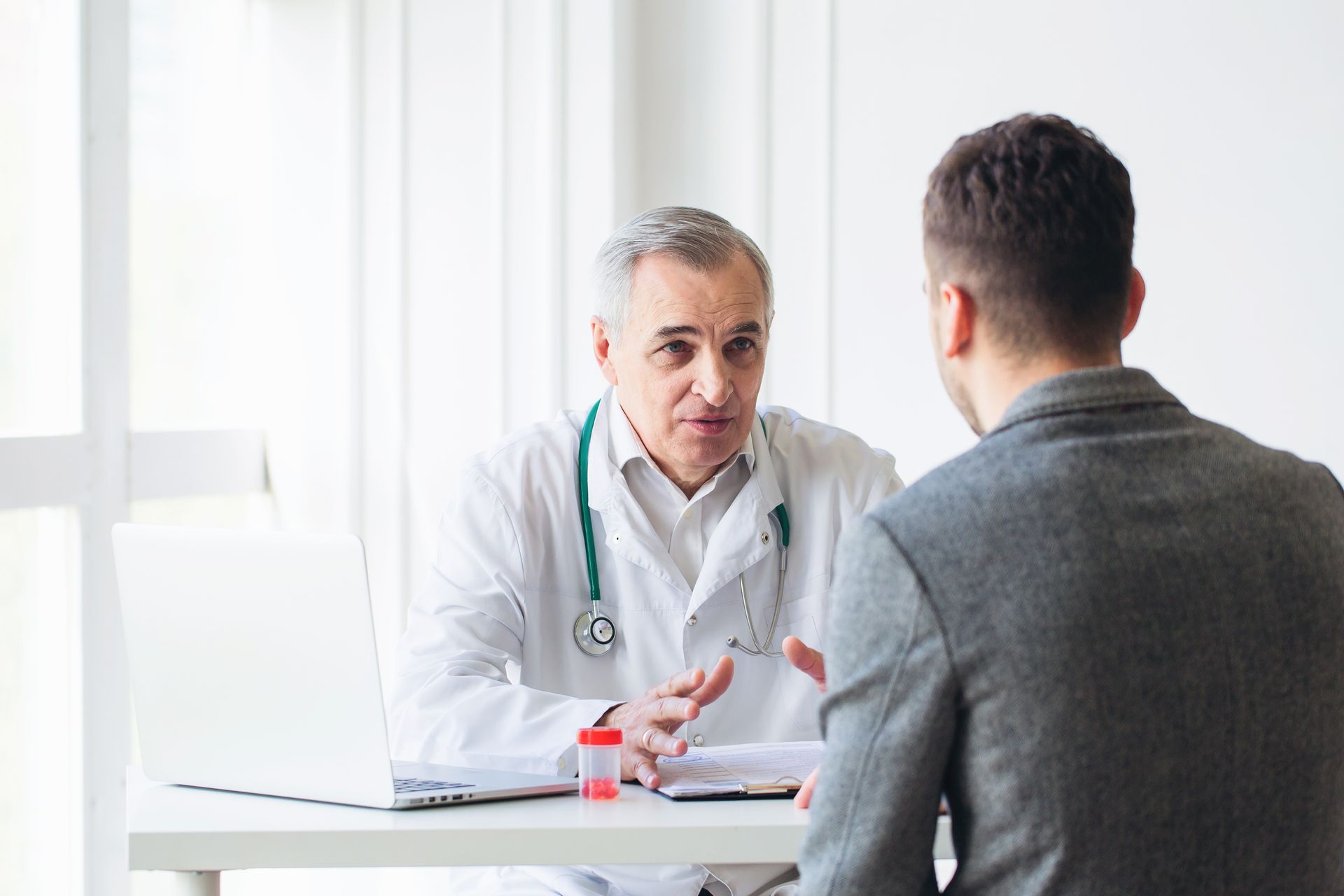 Doctor speaking with a patient in a bright office, seated across a desk with a laptop and stethoscope nearby. Doctor speaking with a patient in a bright office, seated across a desk with a laptop and stethoscope nearby.